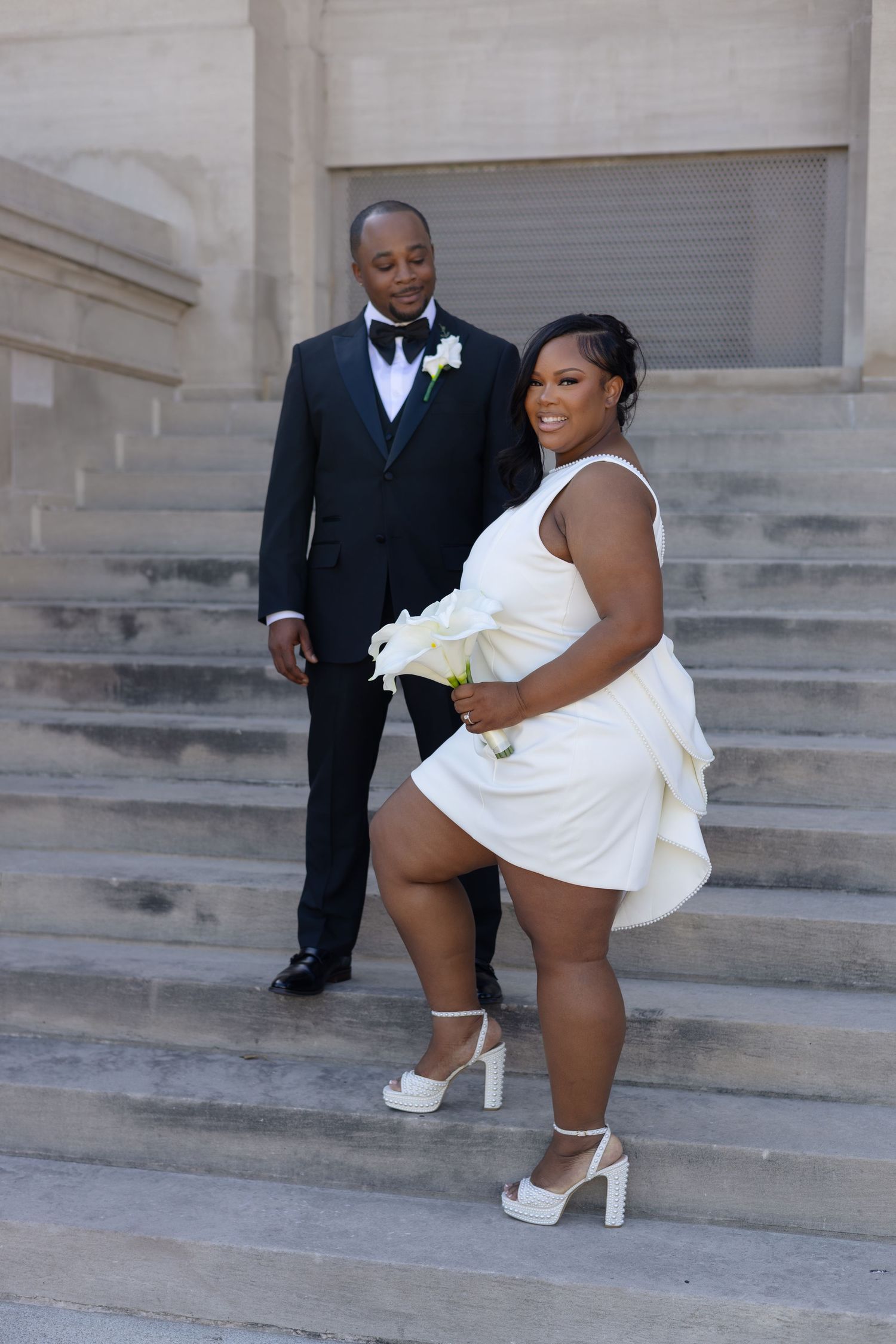 A bride in a short white dress and groom in a black tuxedo pose on stone steps during their wedding day.