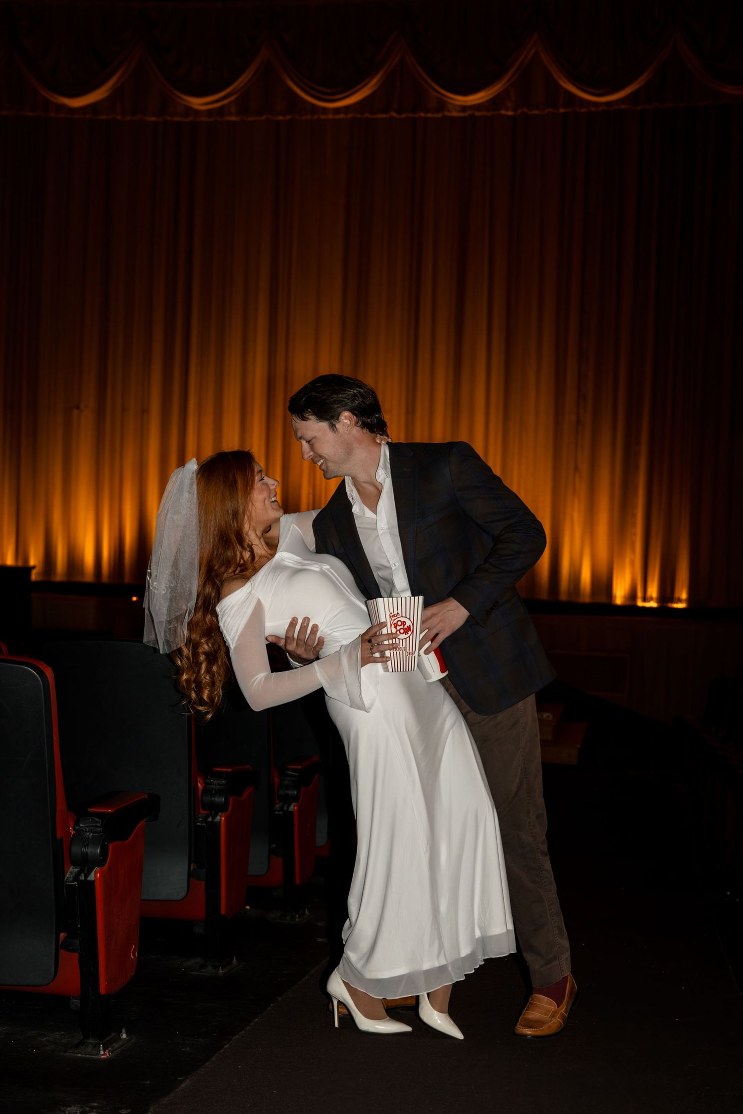 A couple in formal wear shares an intimate moment in front of an illuminated theater curtain.