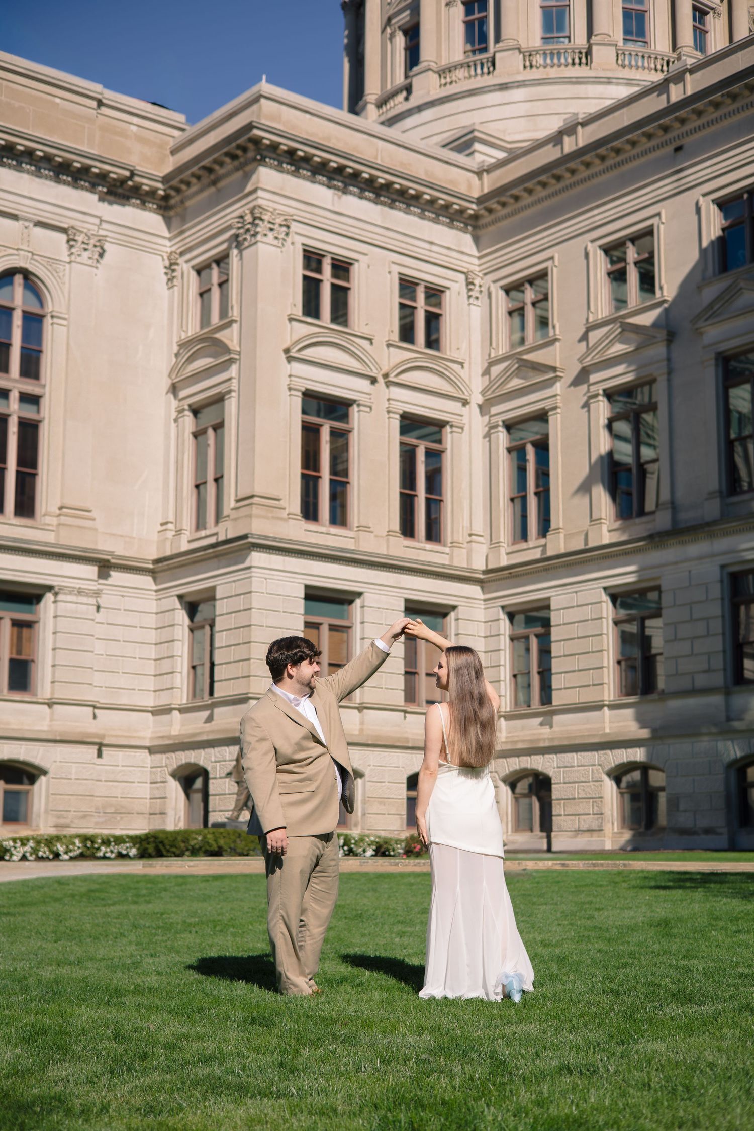 Couple in formal attire shares romantic moments on a grassy lawn outside an elegant historic stone building.