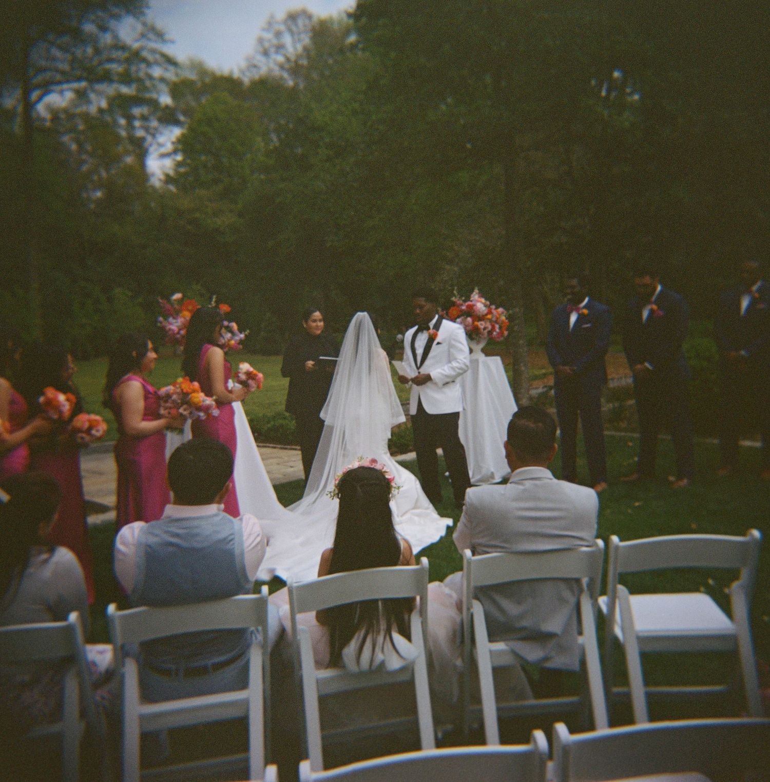 Outdoor wedding ceremony with bride in white gown and veil, groom, bridesmaids in pink, and groomsmen in a lush green park.