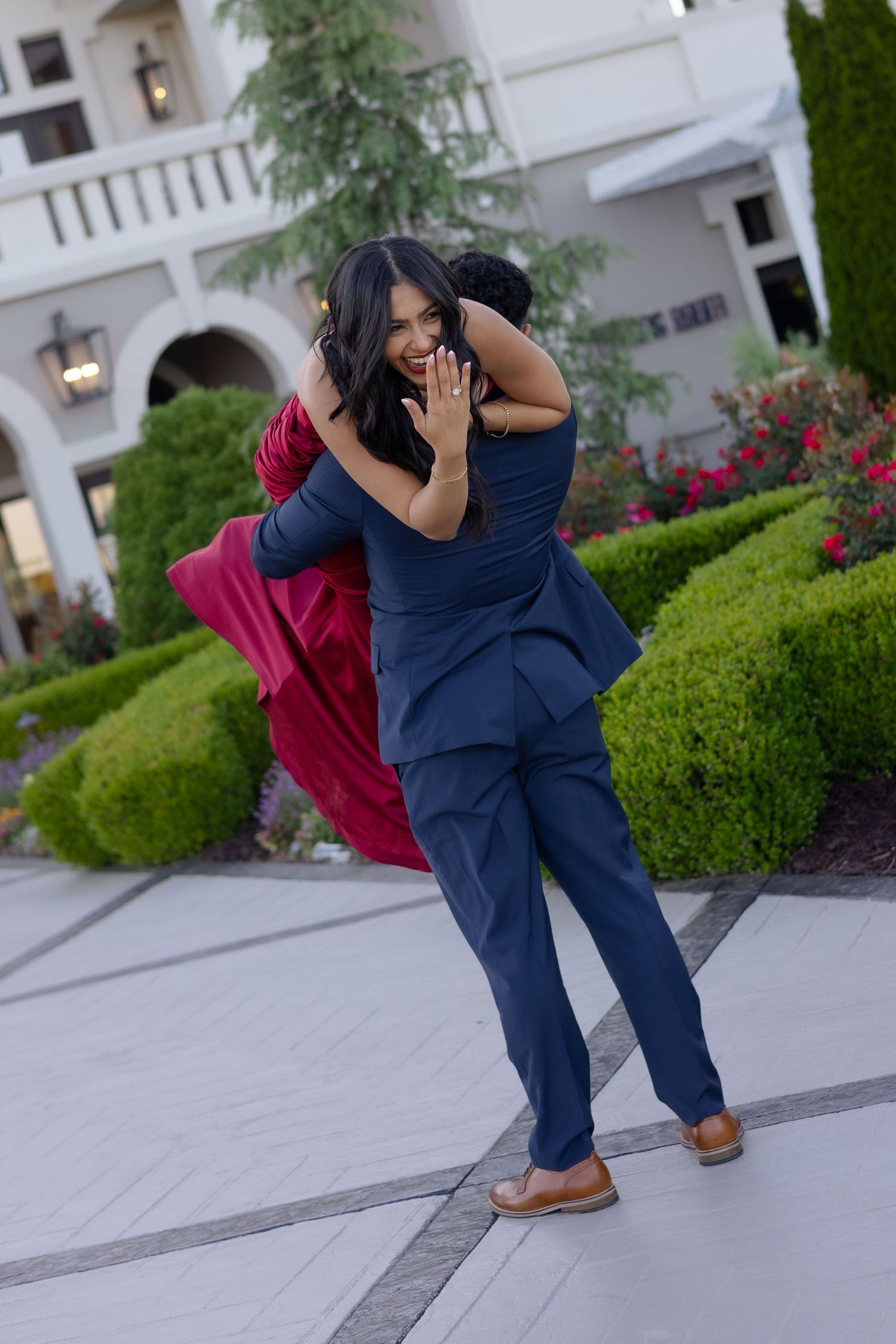 A man in a navy suit lifts a woman in a red dress outdoors near manicured hedges and a white building.