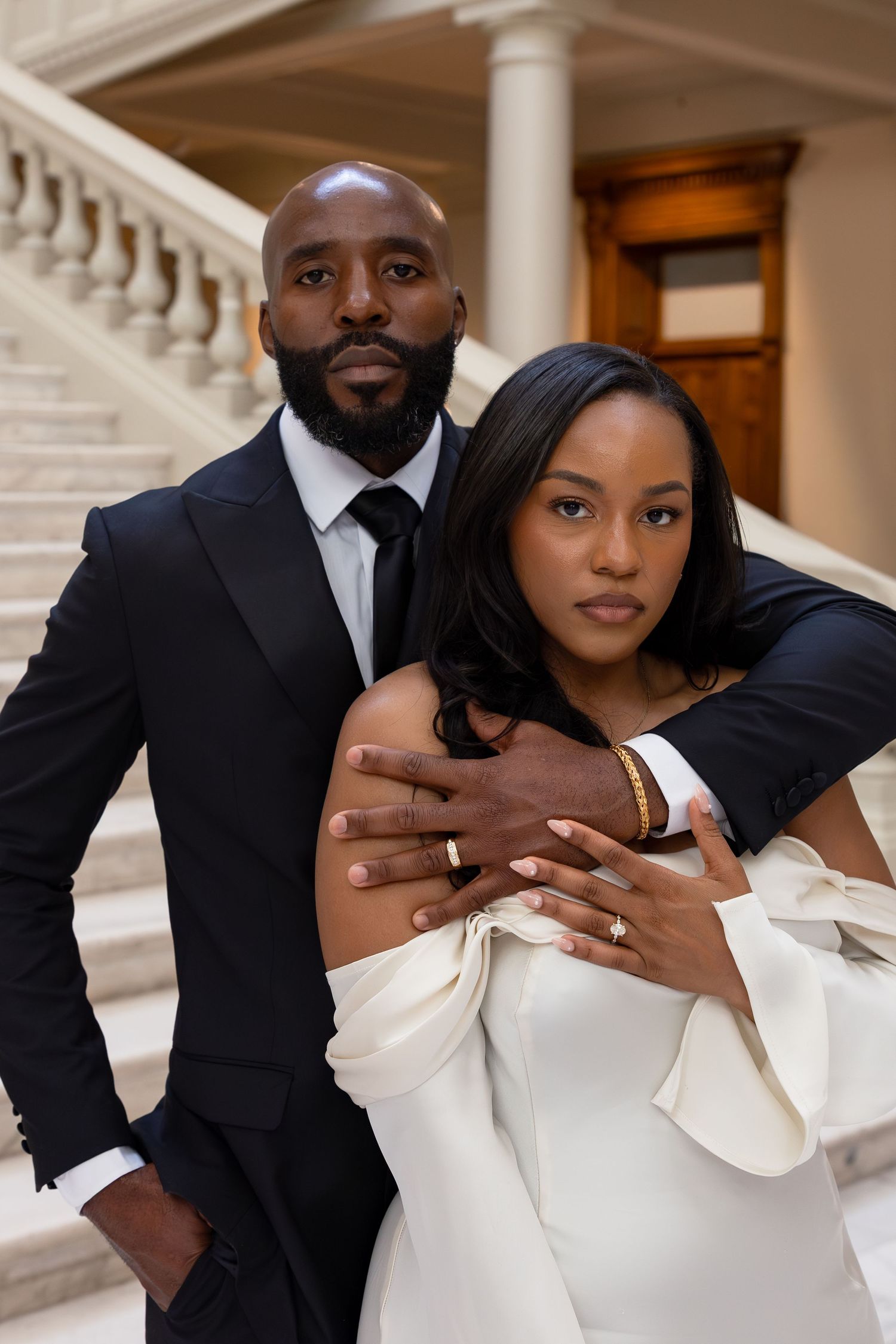 Elegant couple in formal attire posing on marble staircase, man in black suit embracing woman in white off-shoulder gown.