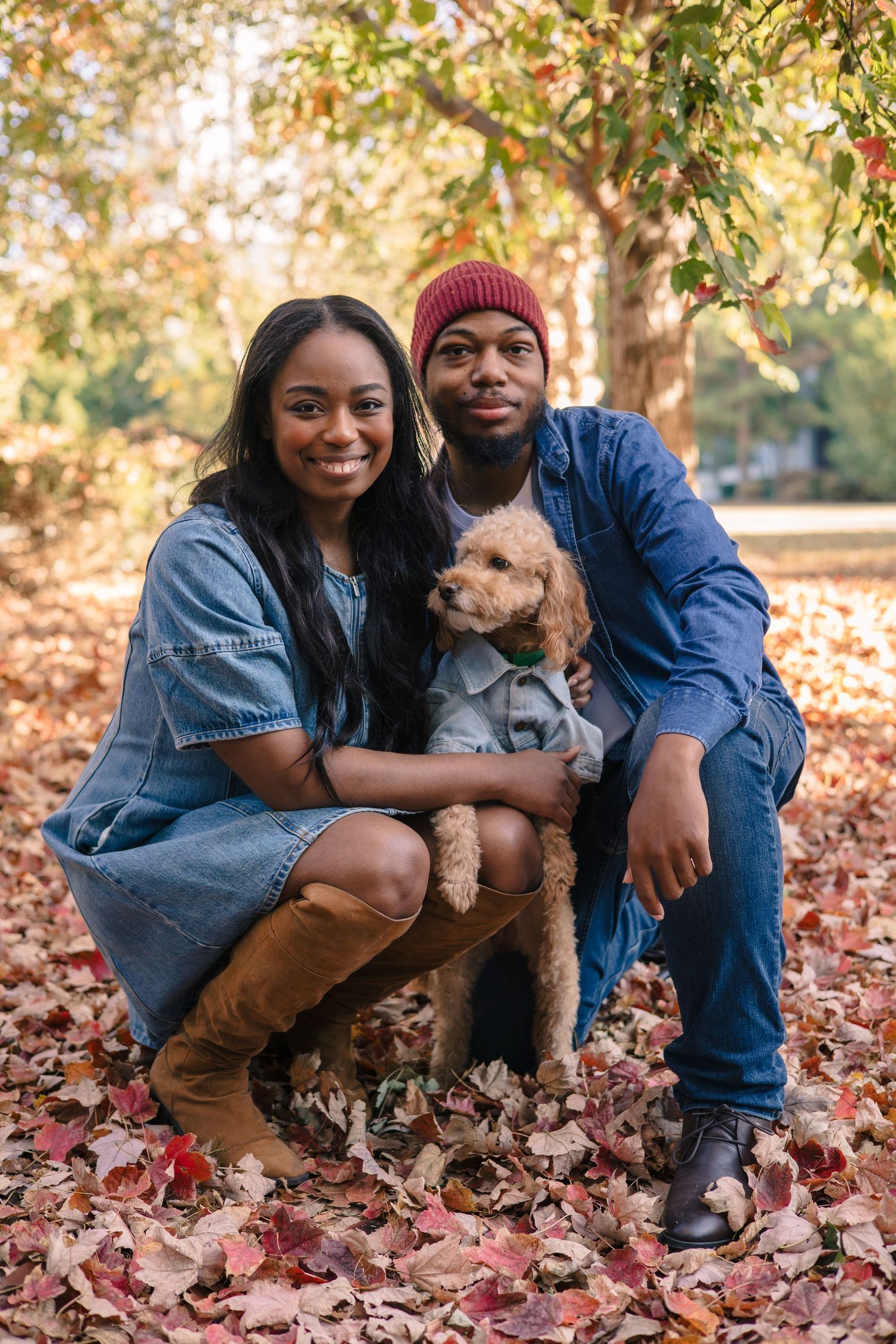 A happy couple sitting in a park surrounded by fallen leaves, holding a small dog and smiling at the camera.