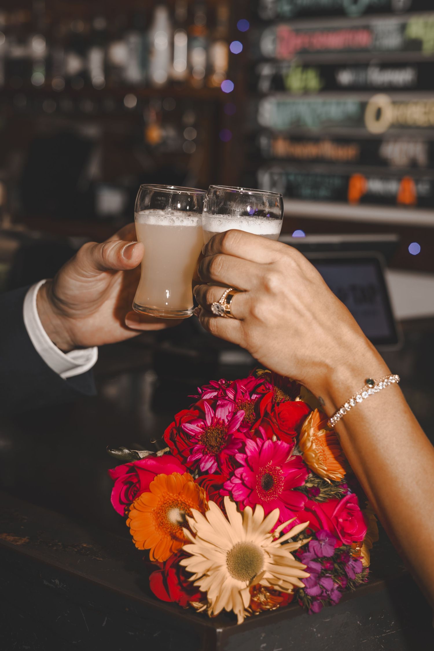 Two hands toast with beer glasses next to a vibrant floral bouquet at a bar.