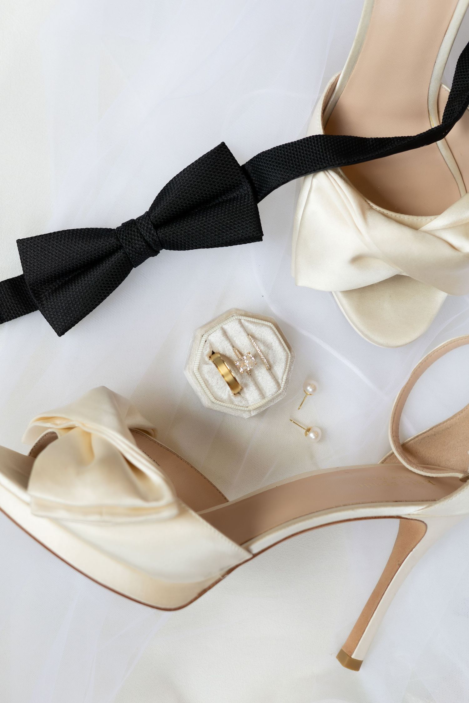 Elegant bridal accessories flat lay with cream satin heels, black bow tie, gold wedding rings, and pearl earrings.