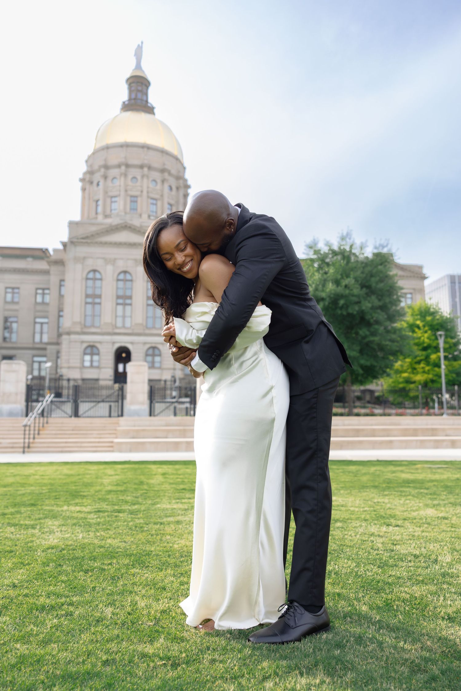 A couple in formal wear embraces on a lawn in front of a grand domed government building on a clear day.