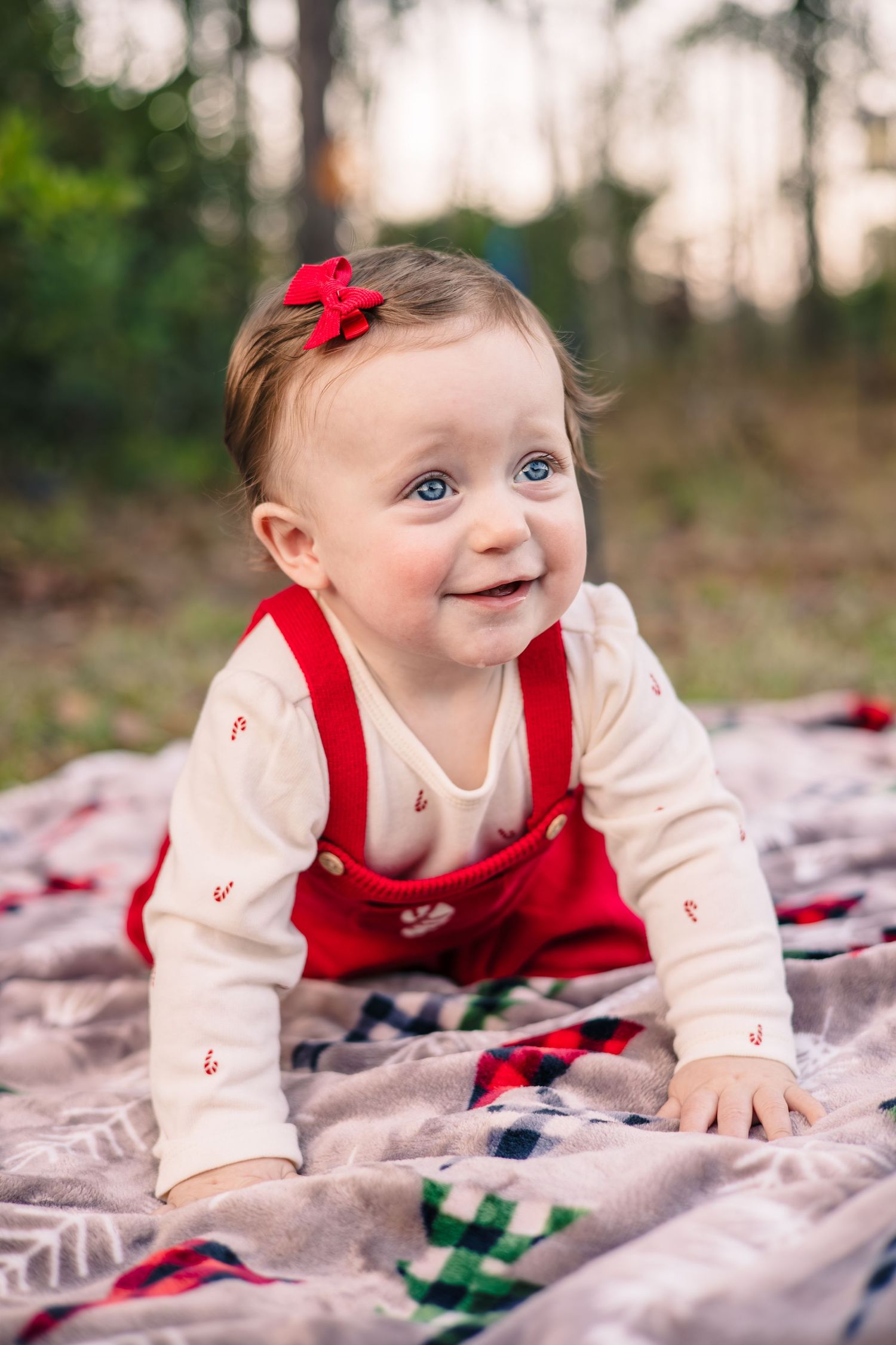 Baby wearing red jumper and white shirt lies on plaid blanket during outdoor photo session.