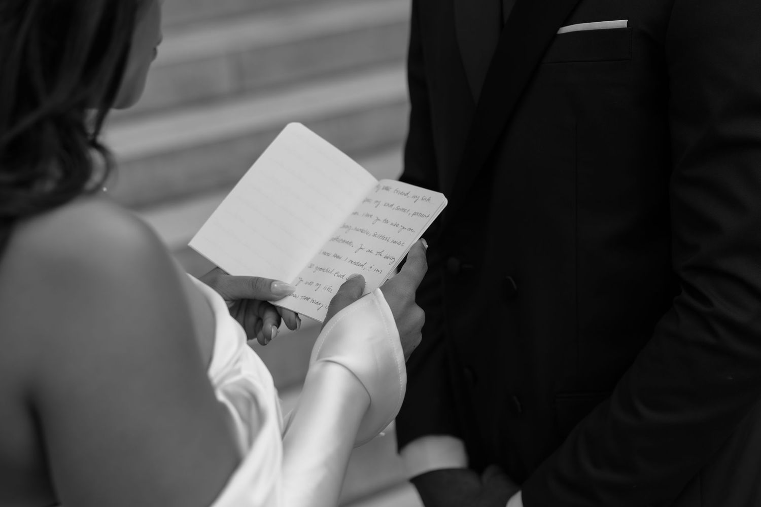 Bride reading wedding vows from a small booklet during an outdoor ceremony in black and white.