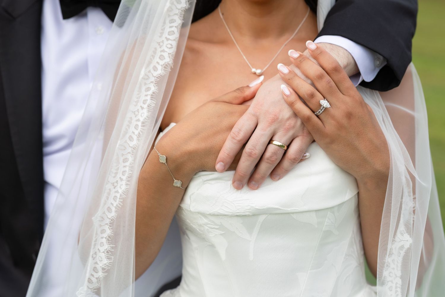 Bride and groom showing wedding rings, bride in white strapless gown with veil, groom in dark suit, close-up of hands.