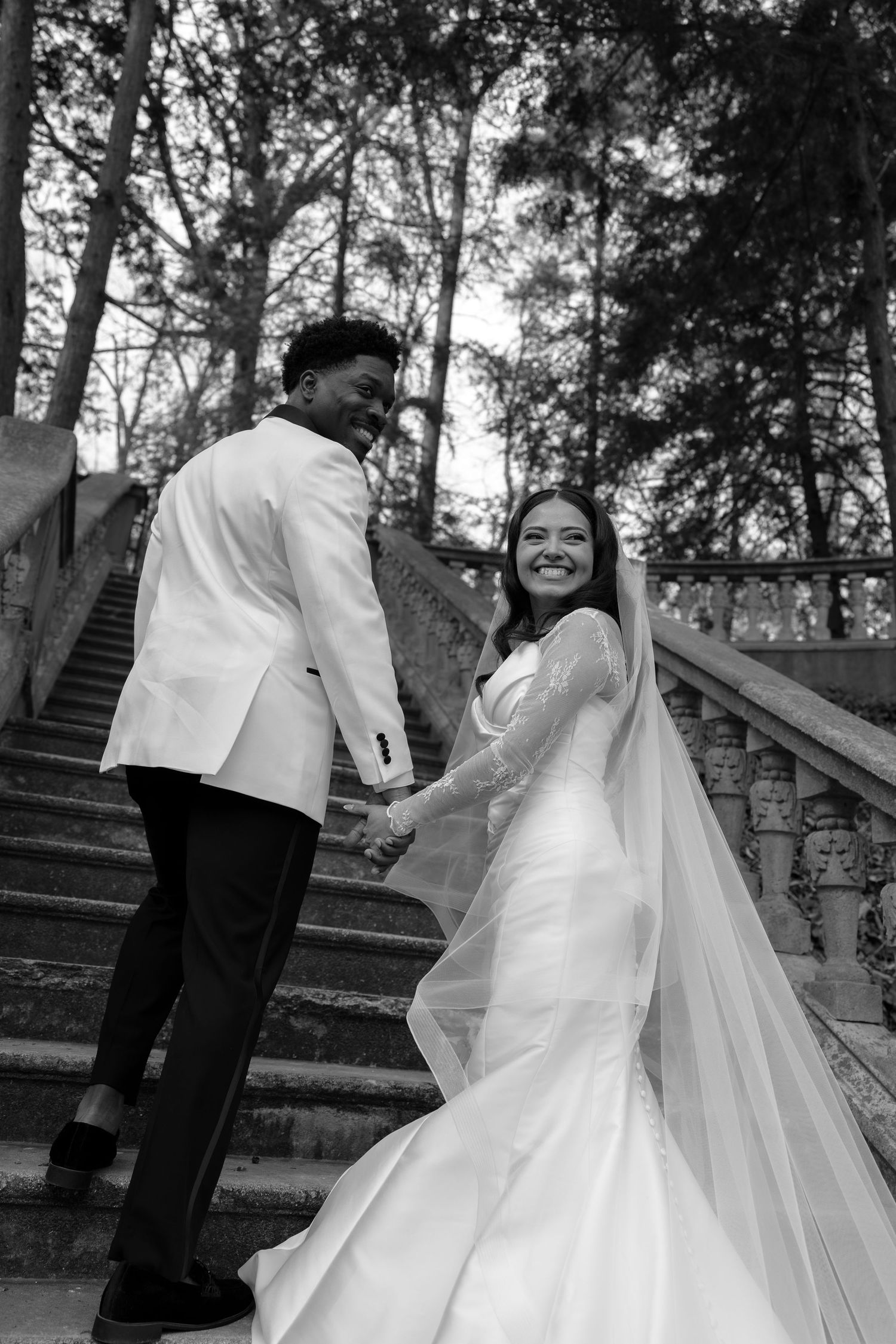 A joyful wedding couple poses on stone steps surrounded by trees in a black and white photograph.