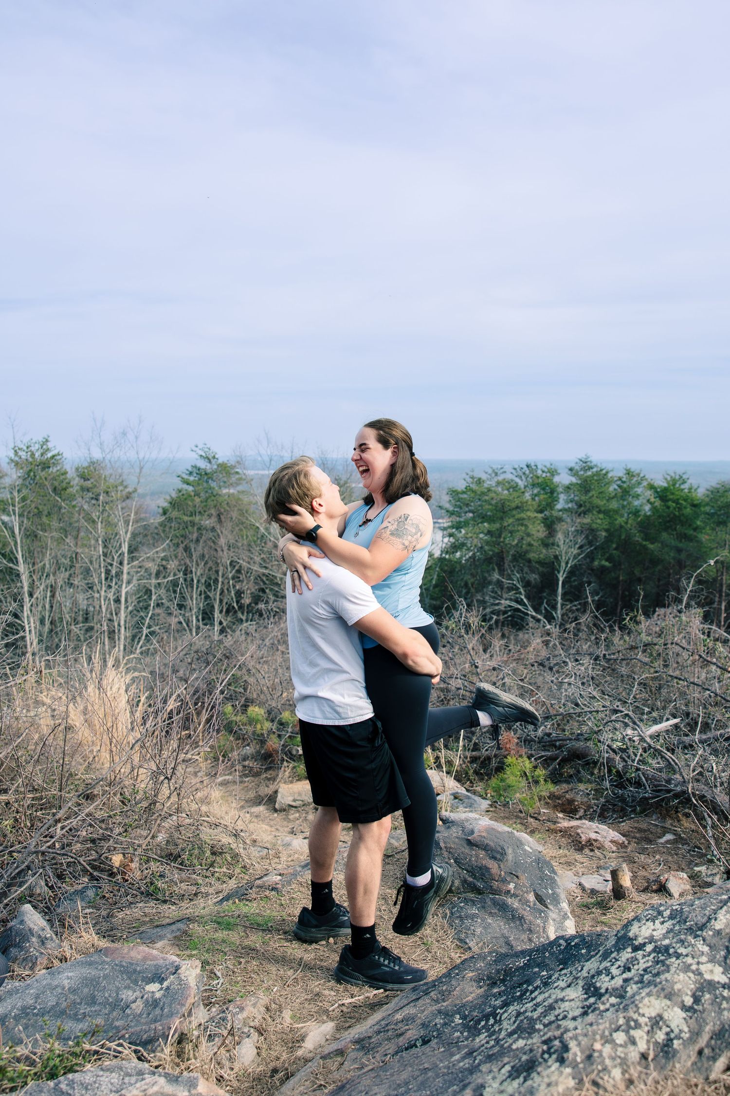 A couple embraces and celebrates on a rocky mountain overlook with trees in the background.