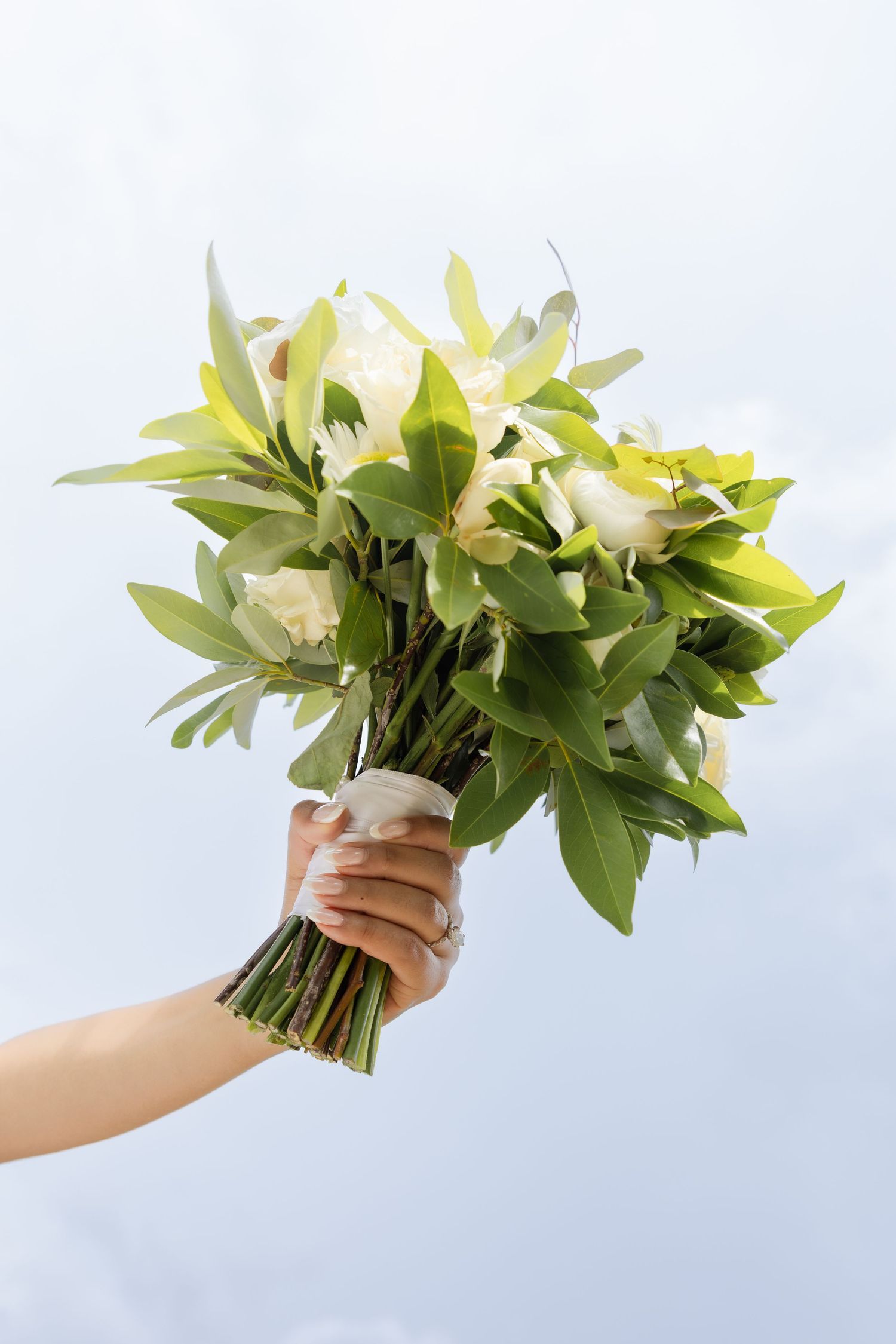 Hand holding a bridal bouquet of white roses and green leaves against a blue sky.