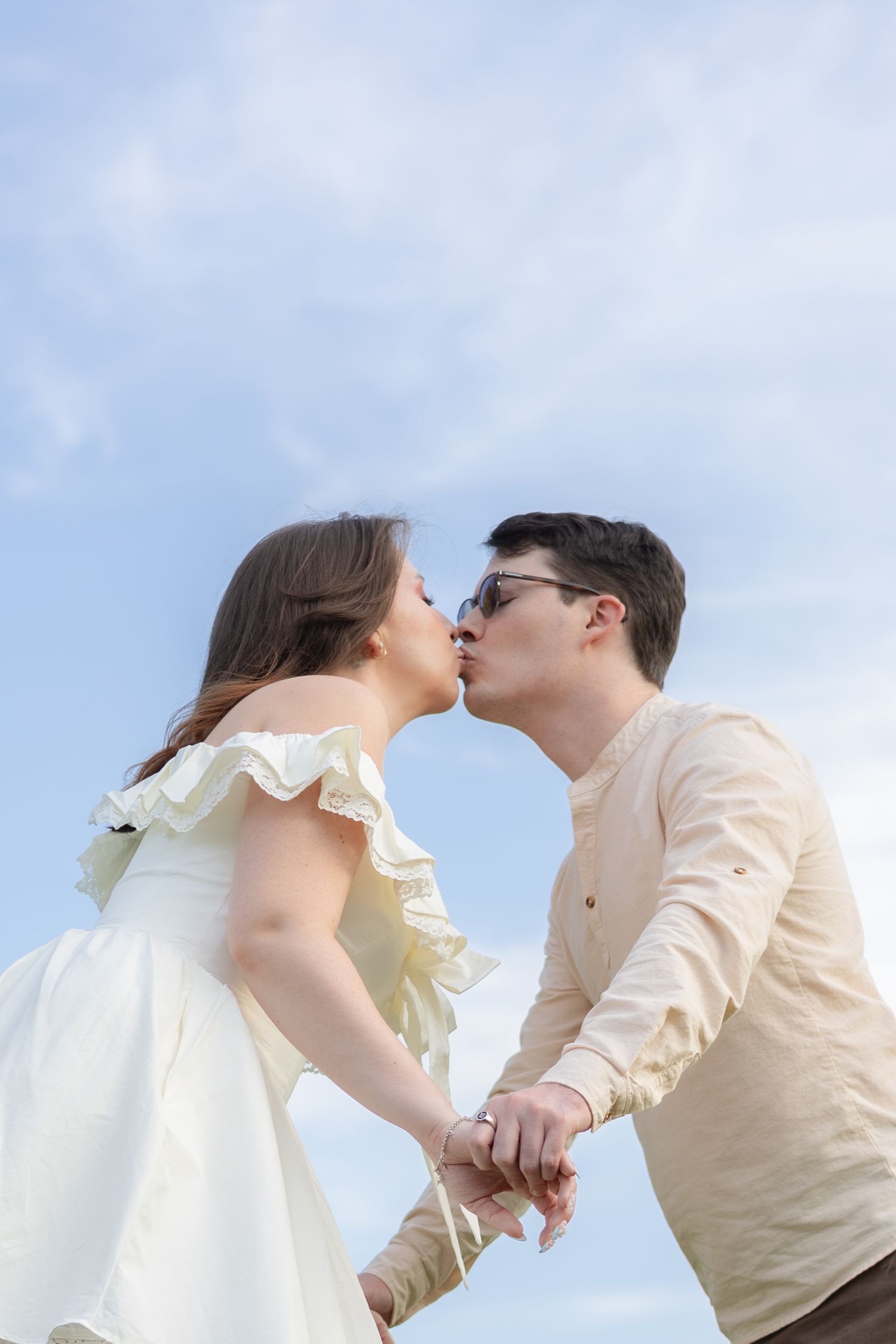 Couple sharing a kiss while holding hands against a bright blue sky backdrop.