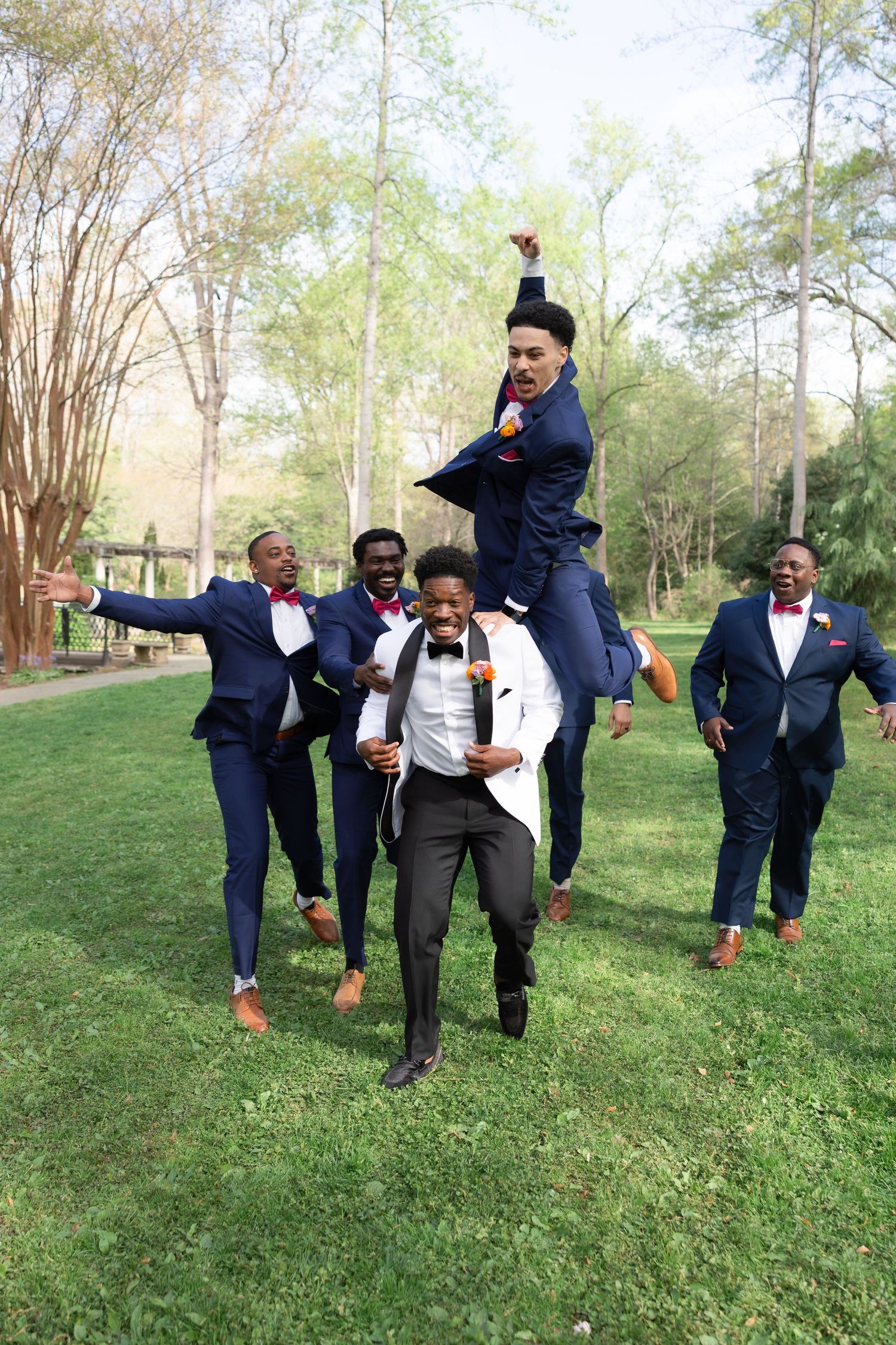 Groomsmen in navy suits with red bow ties celebrate outdoors as one jumps on another's back in a joyful wedding party photo.