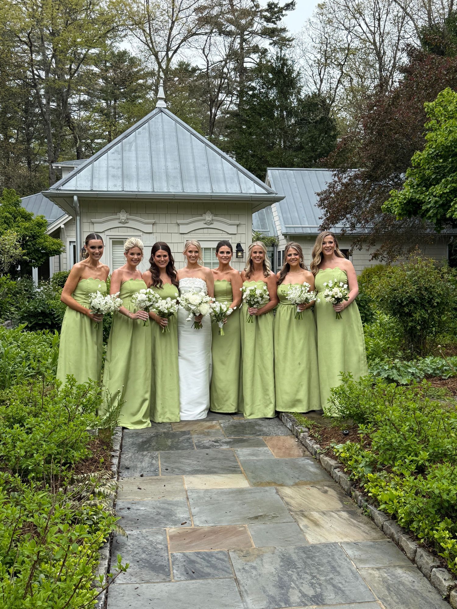 Bridesmaids in light green dresses pose with the bride in white on a stone pathway near a chapel.