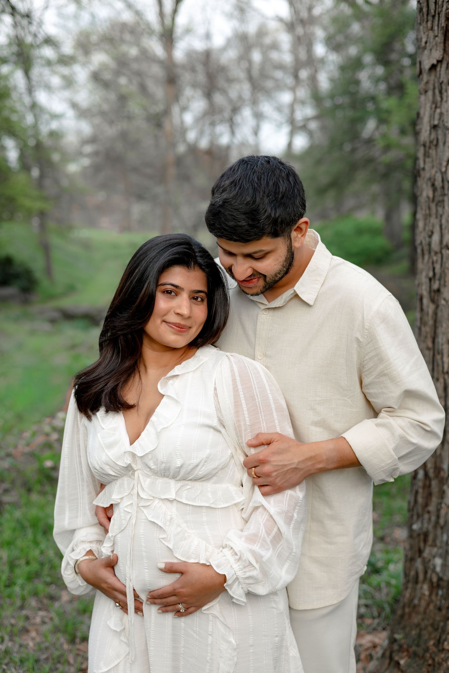 A couple in white clothing embraces tenderly in a wooded outdoor setting during a maternity photo session.