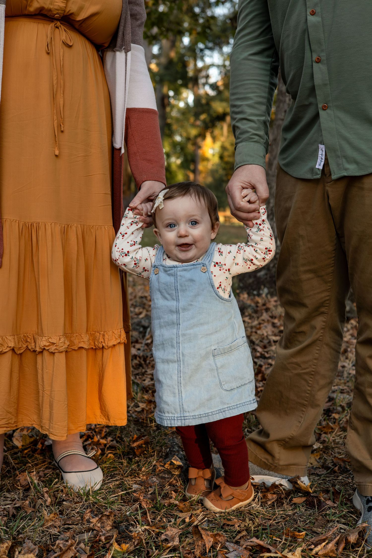 A toddler in denim overalls takes steps on fallen leaves while holding hands with adults.