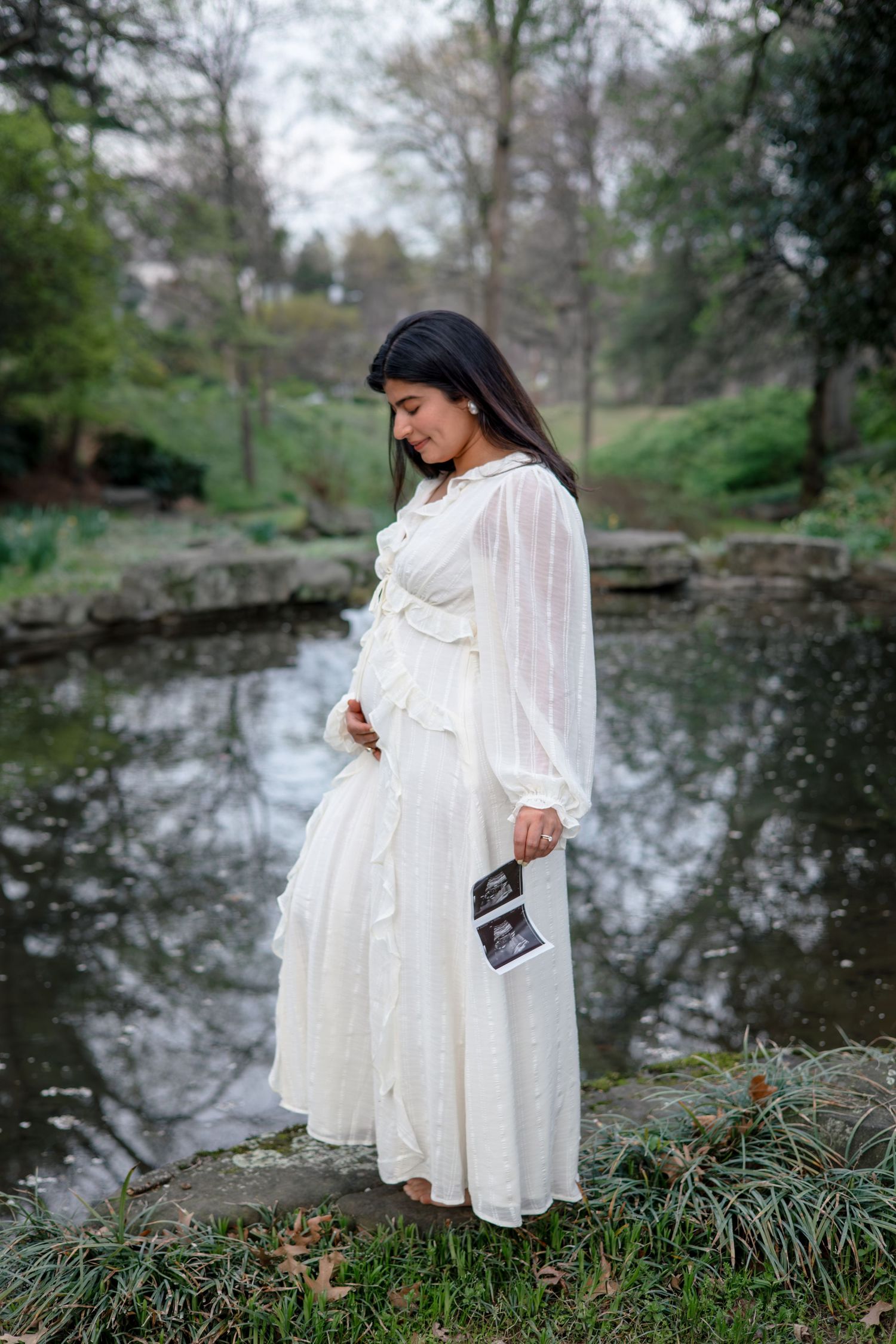 A pregnant woman in a flowing white dress stands by a serene pond while holding ultrasound photos in a natural setting.