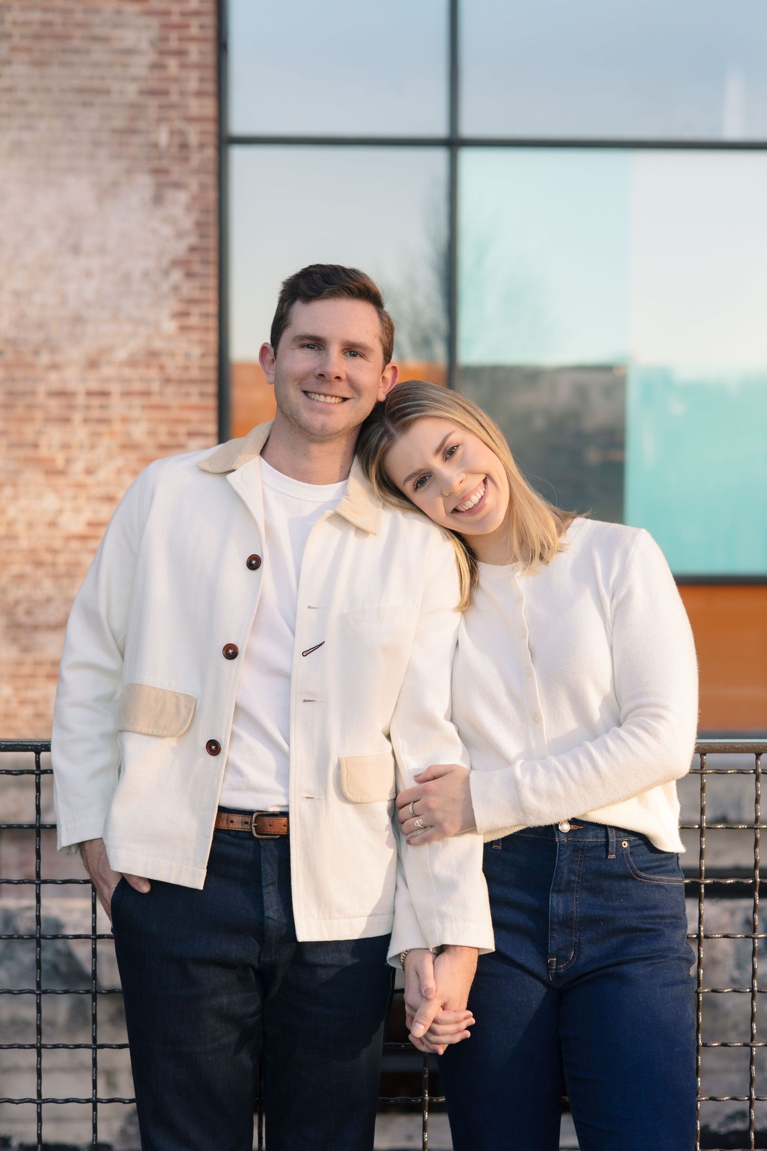 A smiling couple in matching white jackets holds hands outdoors near a brick building.