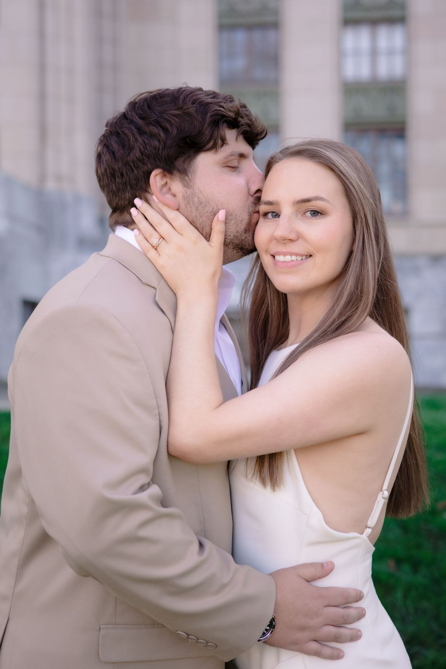 A romantic couple embraces outdoors, with the woman in white and the man in beige sharing an intimate moment.