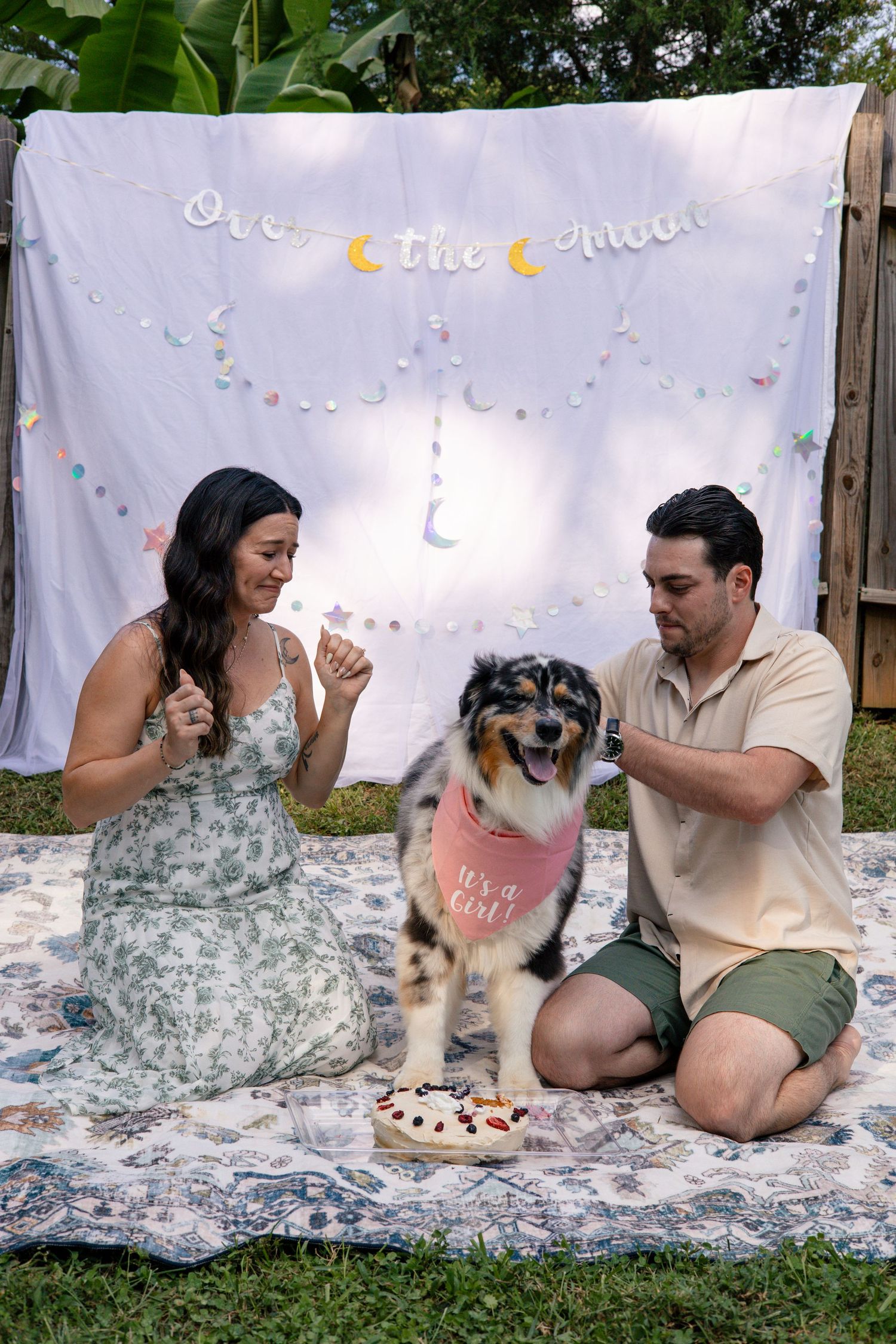 A couple and their dog share cake while sitting on a decorated white blanket outdoors.
