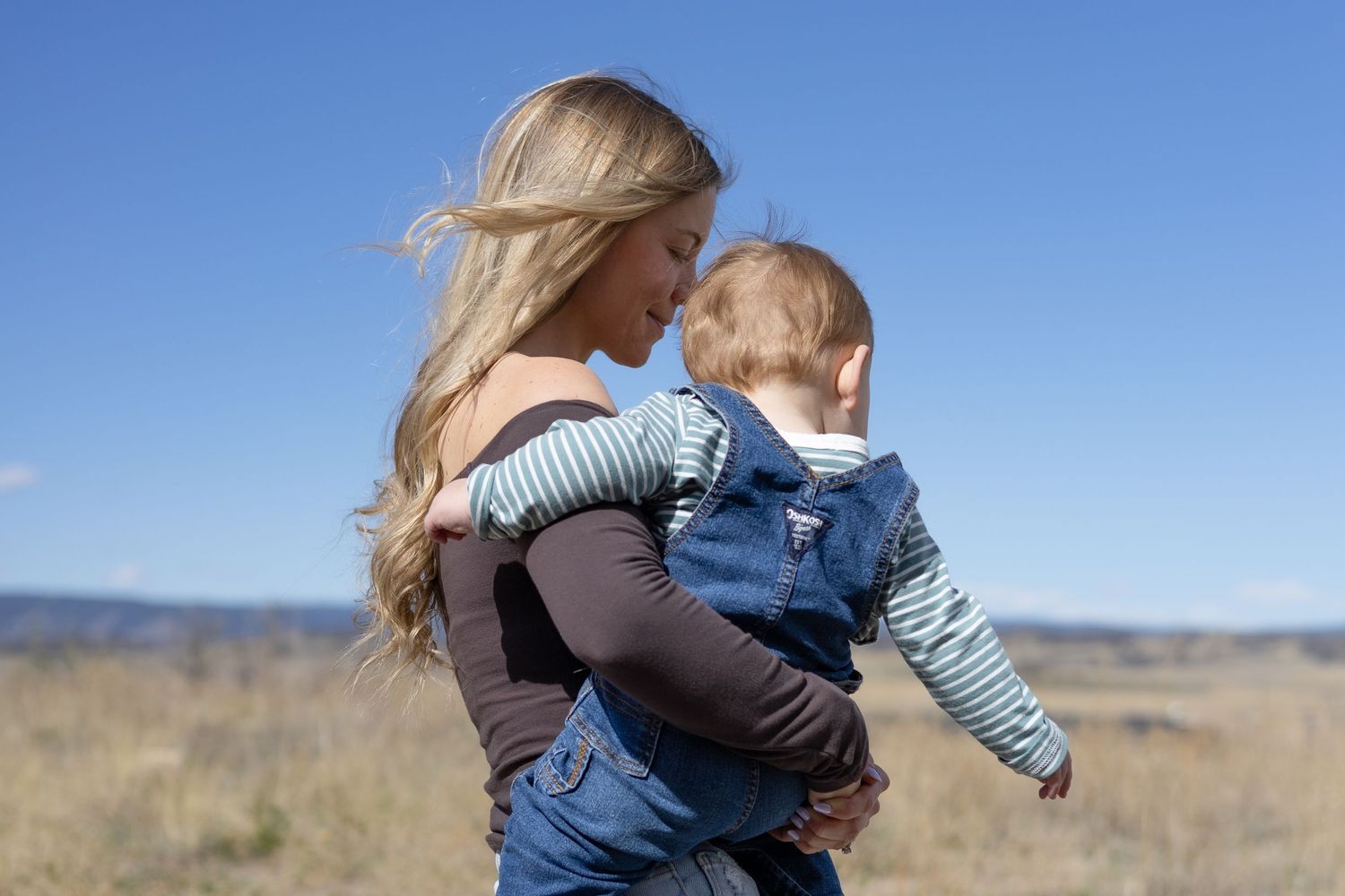 Blonde mother holding baby outdoors in open field under blue sky, side profile view.