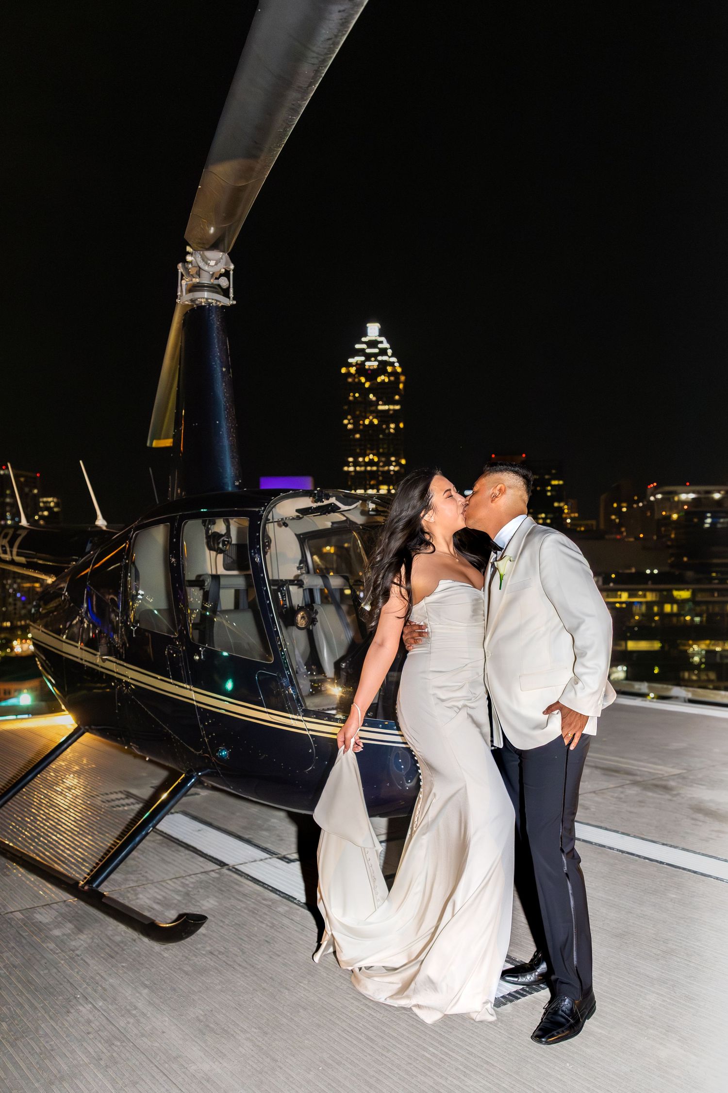 Bride and groom kissing beside helicopter on rooftop at night with Atlanta skyline.