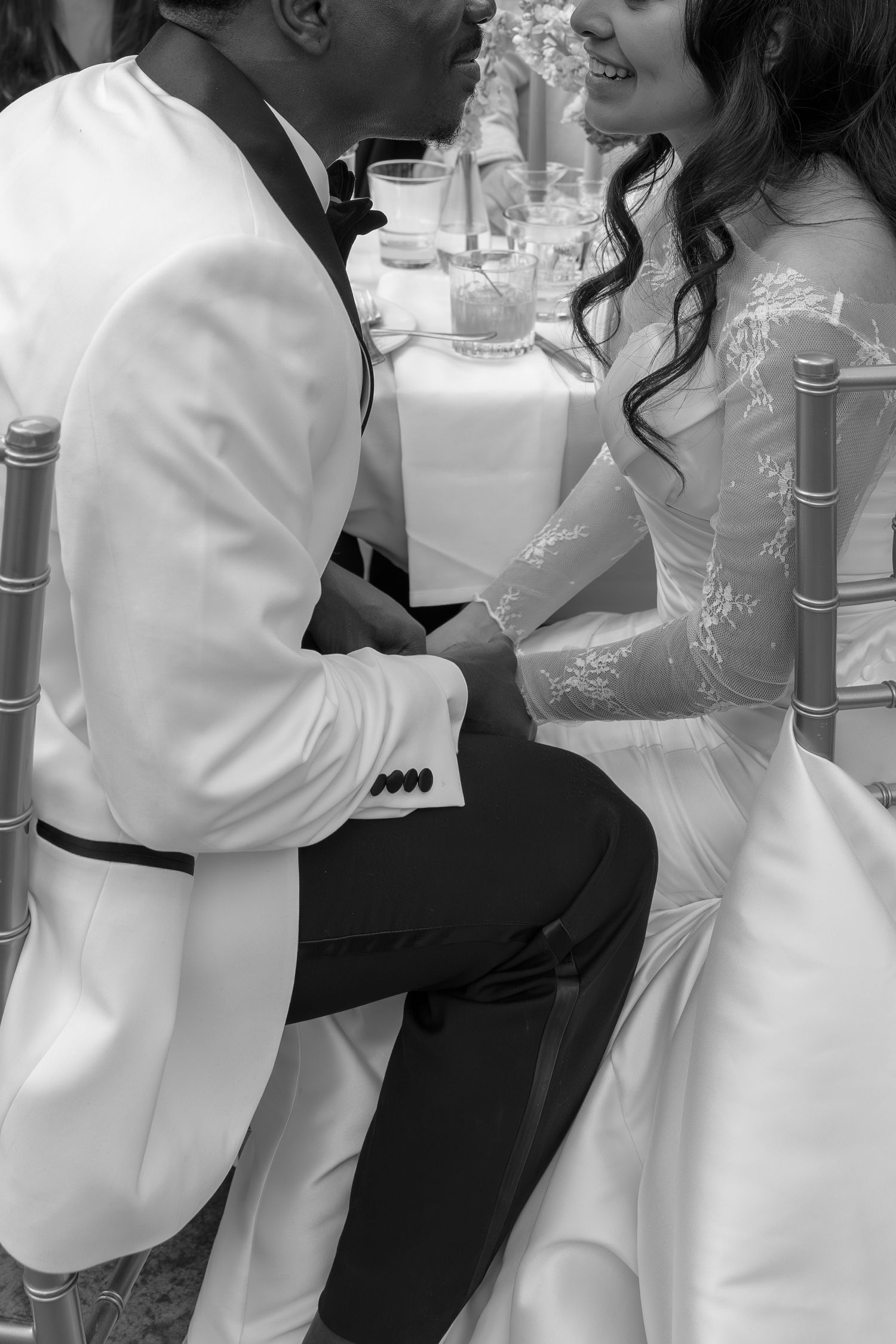 Black and white photo of a bride and groom holding hands and smiling at each other while seated at their wedding reception.