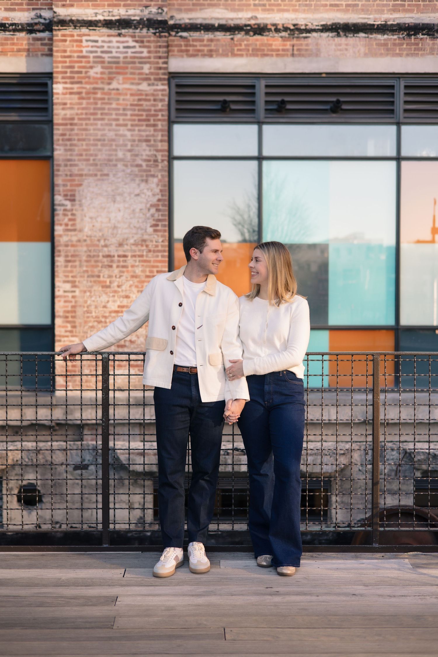 Couple in white shirts posing playfully against brick building exterior.