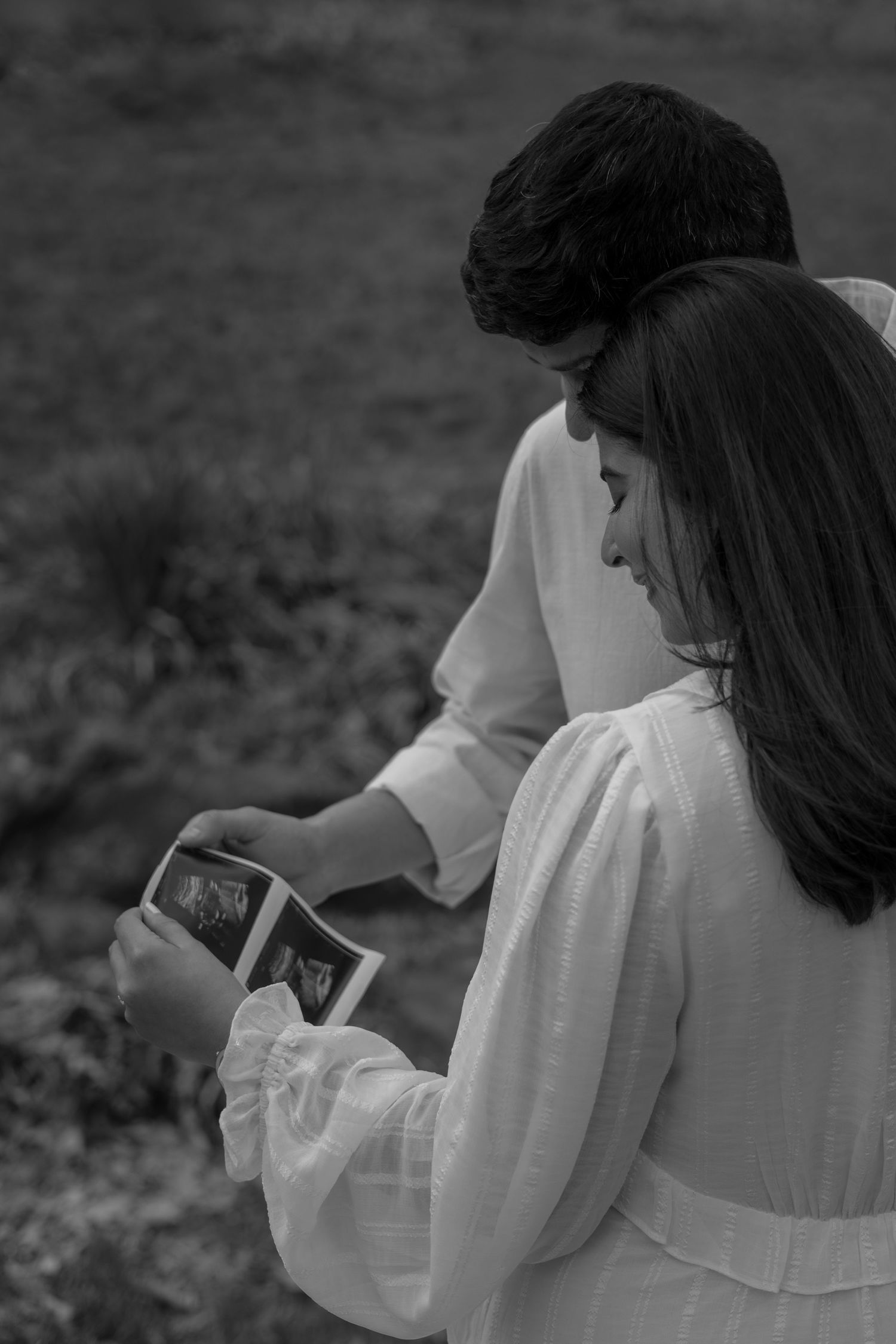 A couple in white clothing sitting outdoors while looking at a sonogram photo together in black and white.