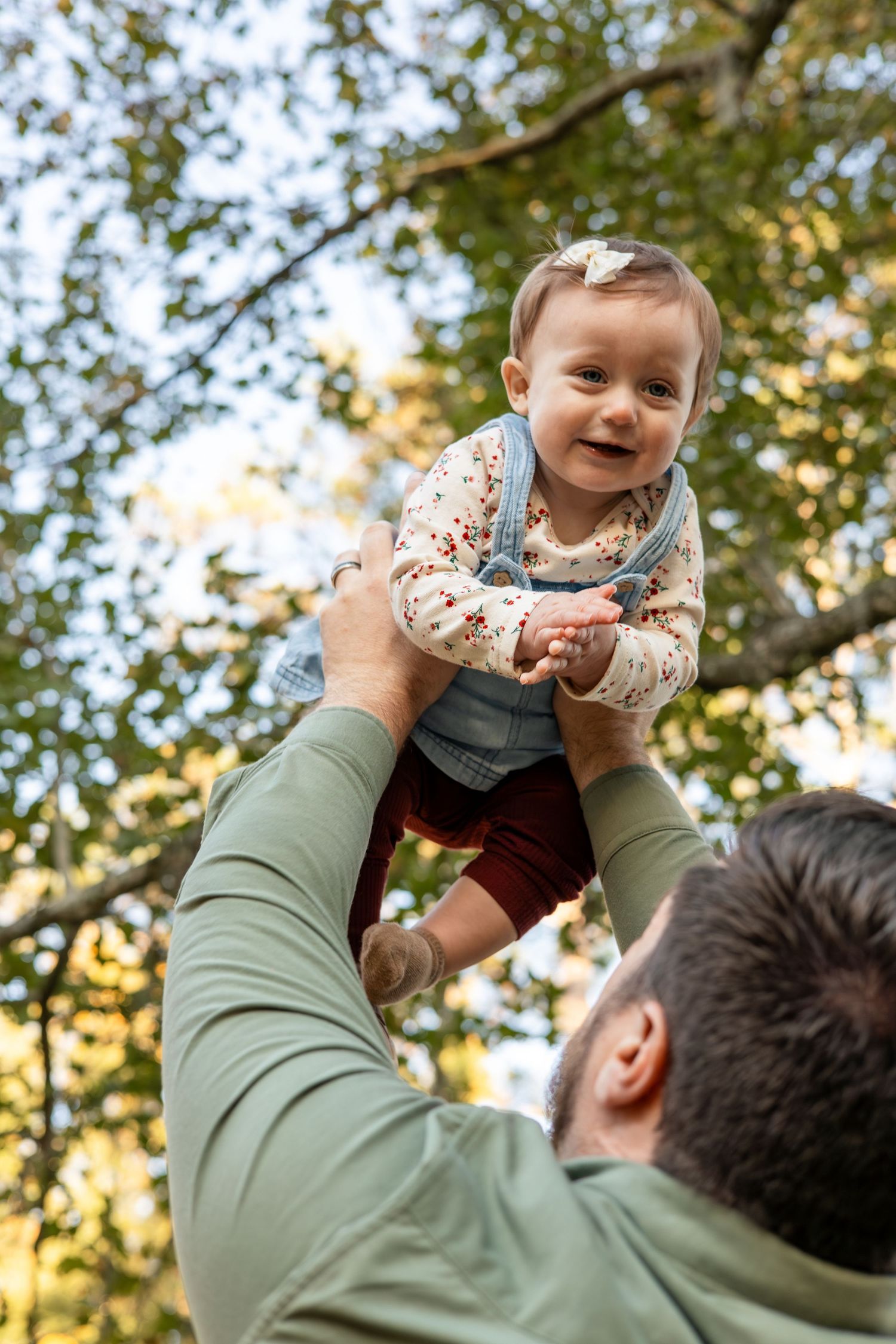 Playful moment captured outdoors with baby being lifted against autumn sky.