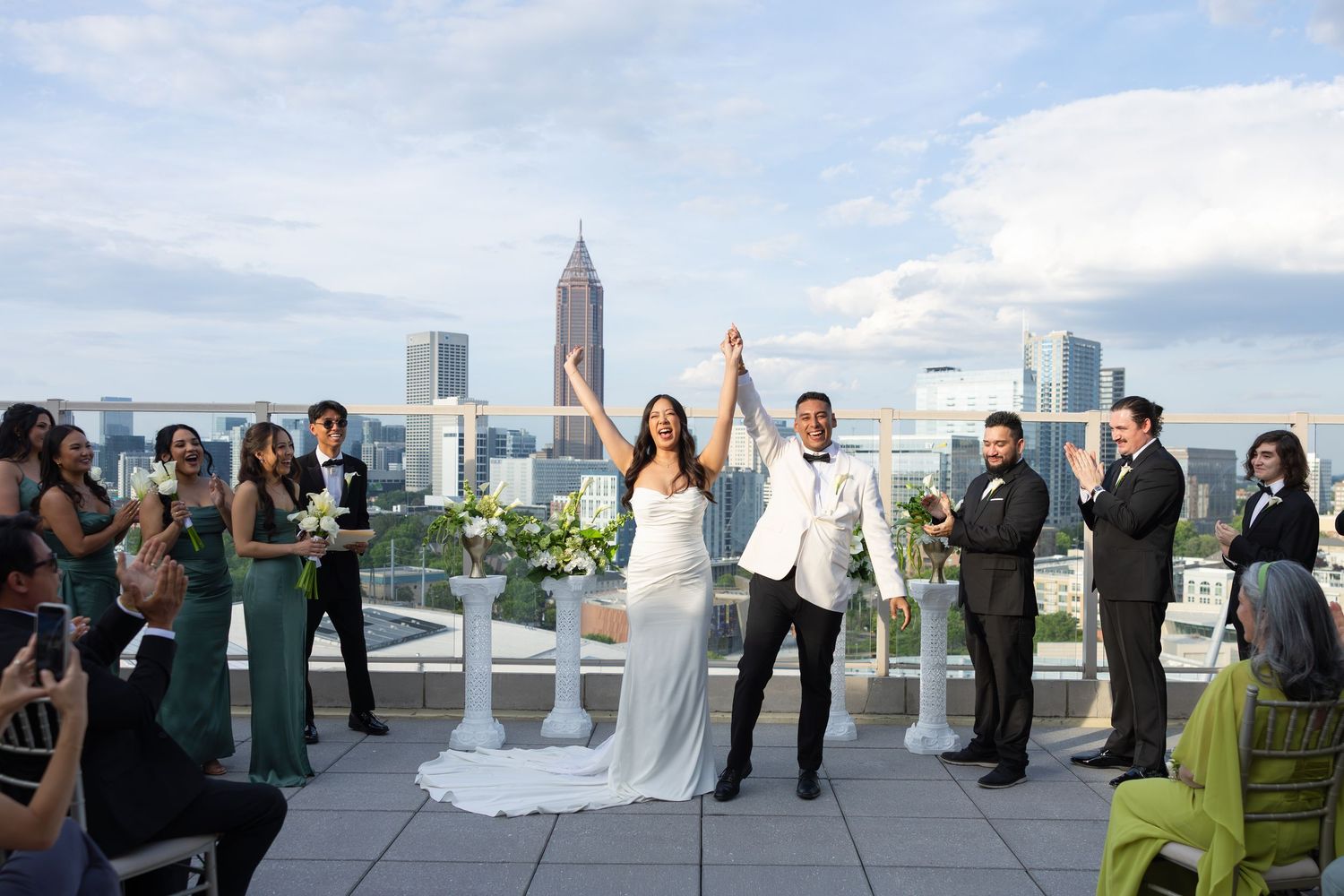 Newlyweds celebrate with raised hands on a rooftop venue with Atlanta city skyline backdrop, guests applauding around them.