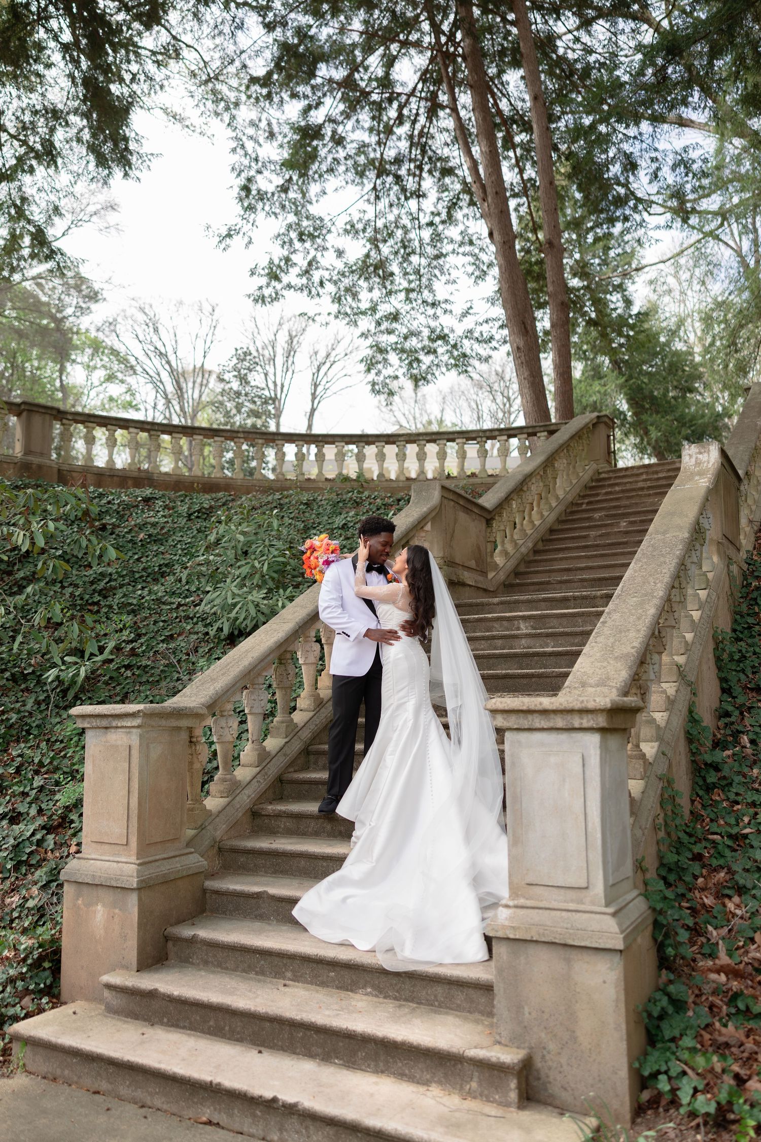 Bride and groom share a romantic moment on elegant stone stairs surrounded by lush greenery on their wedding day.