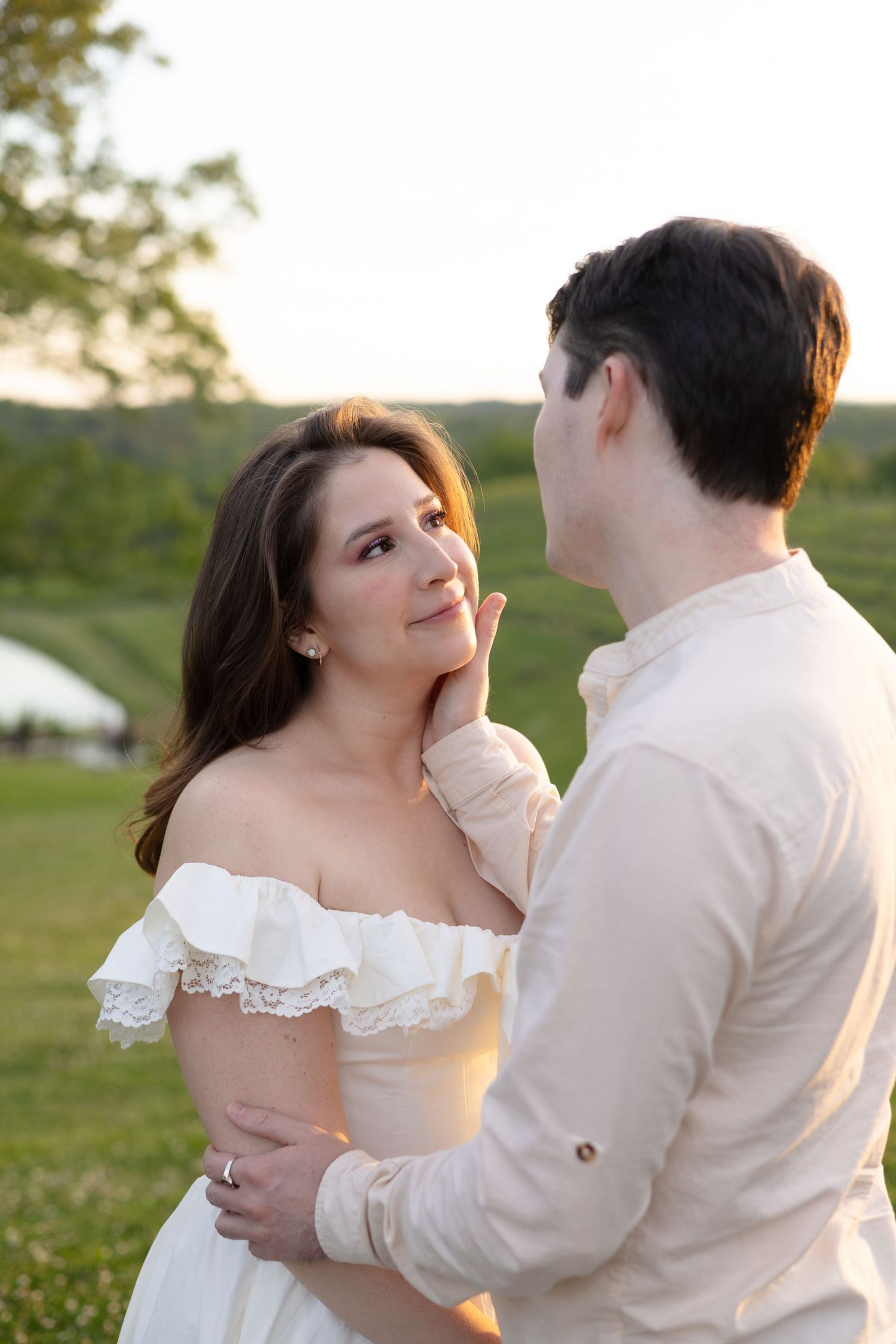 Romantic couple gazing at each other at sunset in a vineyard, woman in white lace off-shoulder dress.