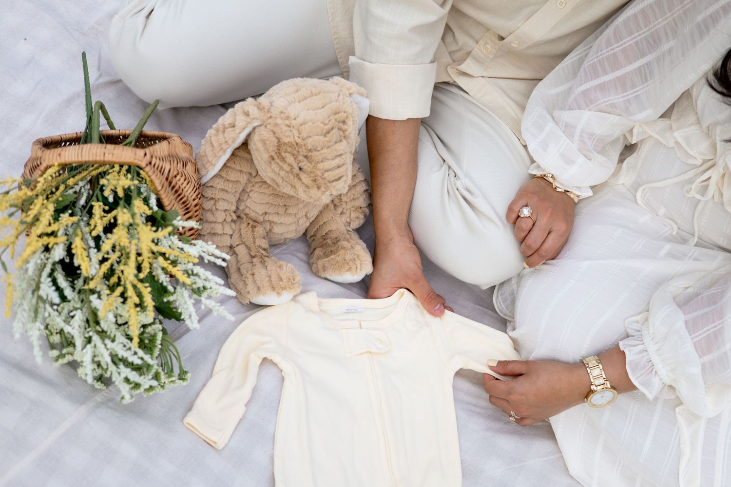 Small white baby clothes and teddy bear arranged on white bedding next to a bouquet of yellow and white flowers.