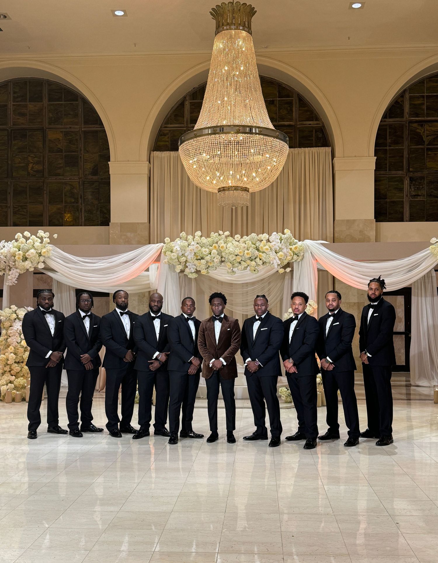 Groomsmen in black tuxedos pose in an elegant ballroom with crystal chandeliers and white drapery.