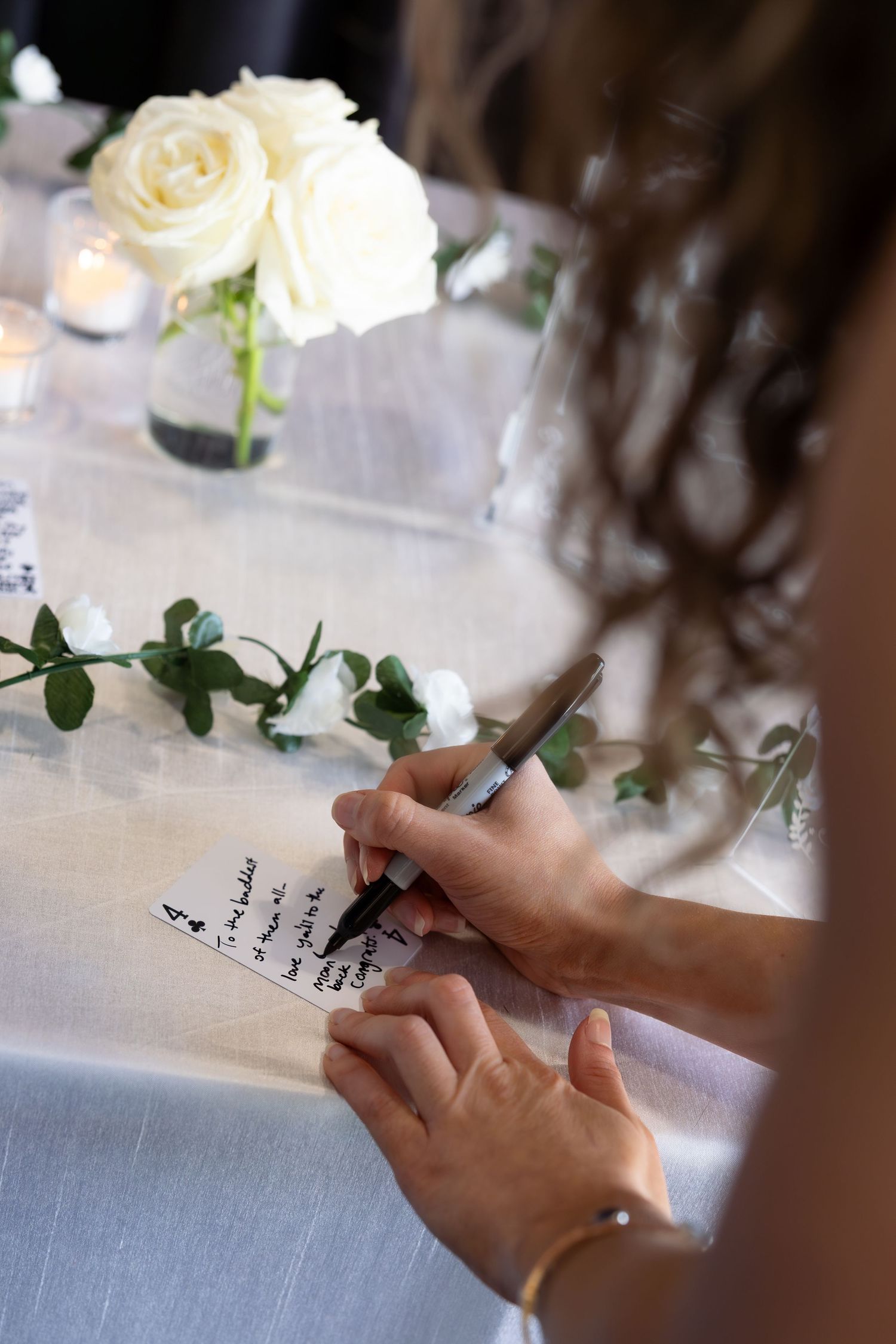 Bride signing wedding guest book at decorated table with white roses and candles.