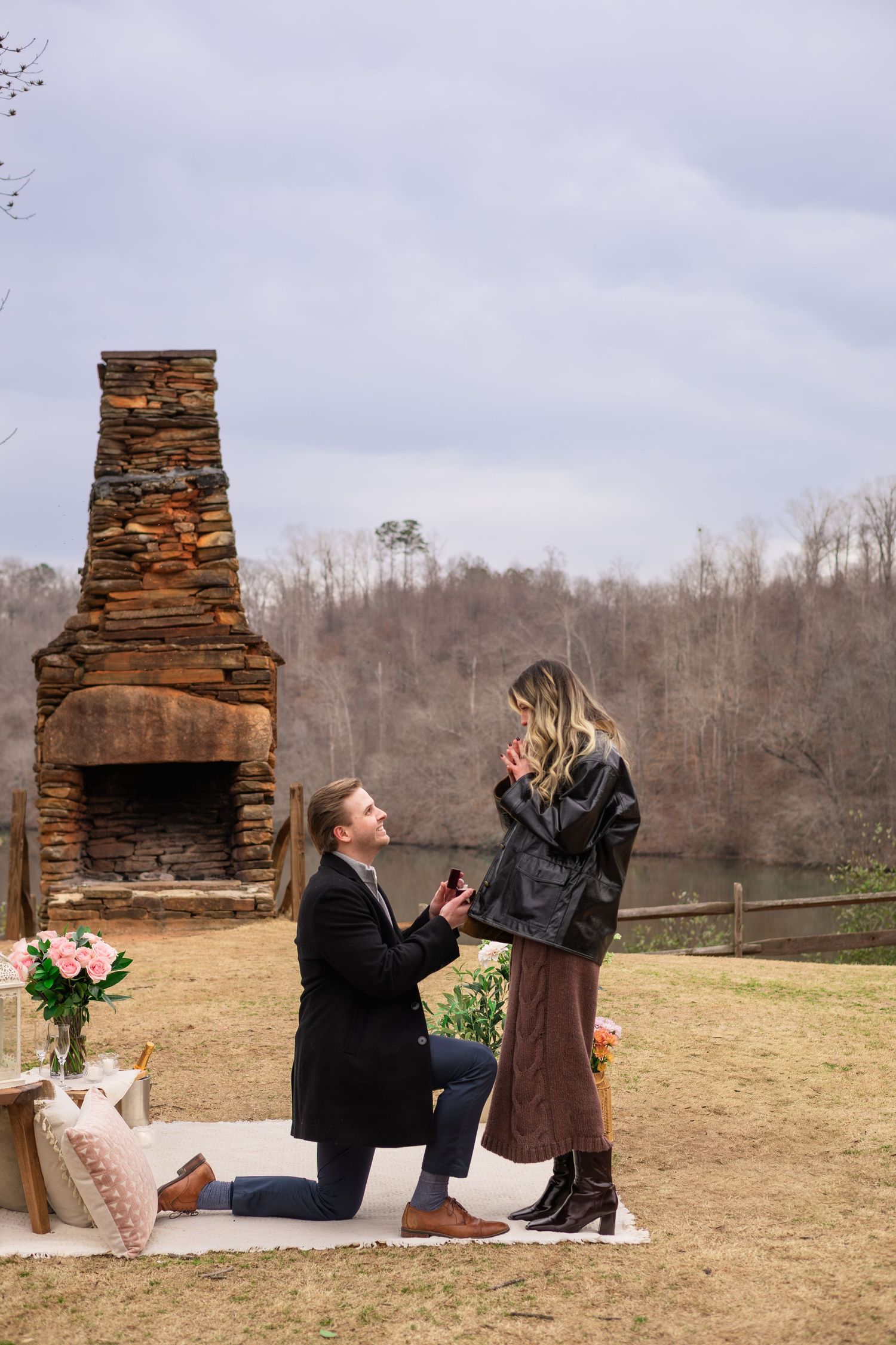 A romantic proposal scene unfolds in front of a rustic stone chimney on a grassy hillside.