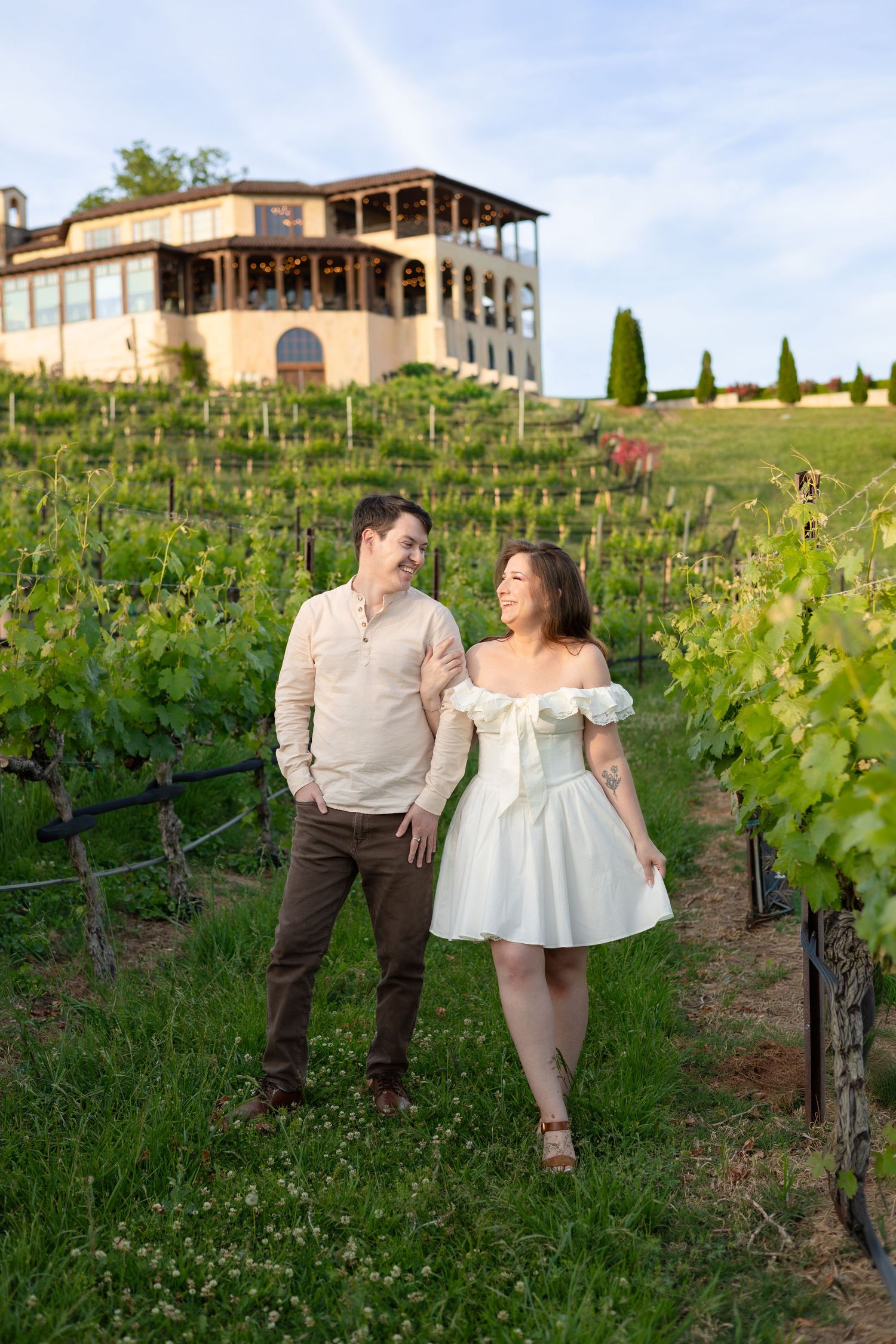 Couple holding hands in a lush vineyard with a winery building on a hill, woman in white dress, man in beige shirt.