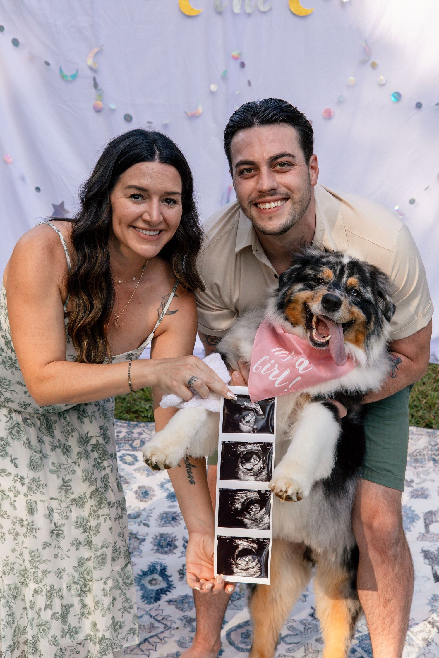A couple poses with their dog and ultrasound photo on a decorated white blanket.