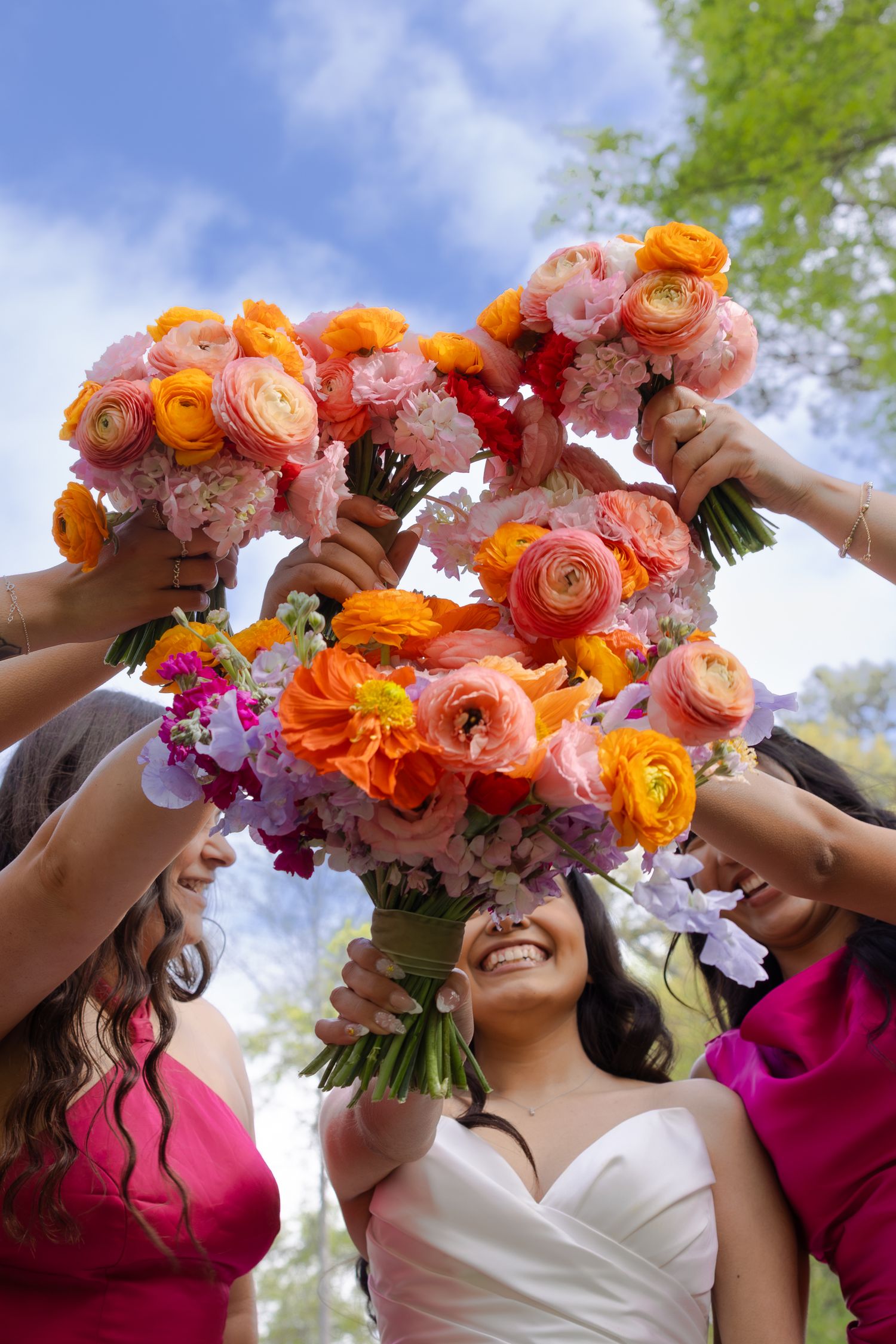 Bride and bridesmaids in pink dresses raise colorful orange and pink floral bouquets outdoors under a blue sky.