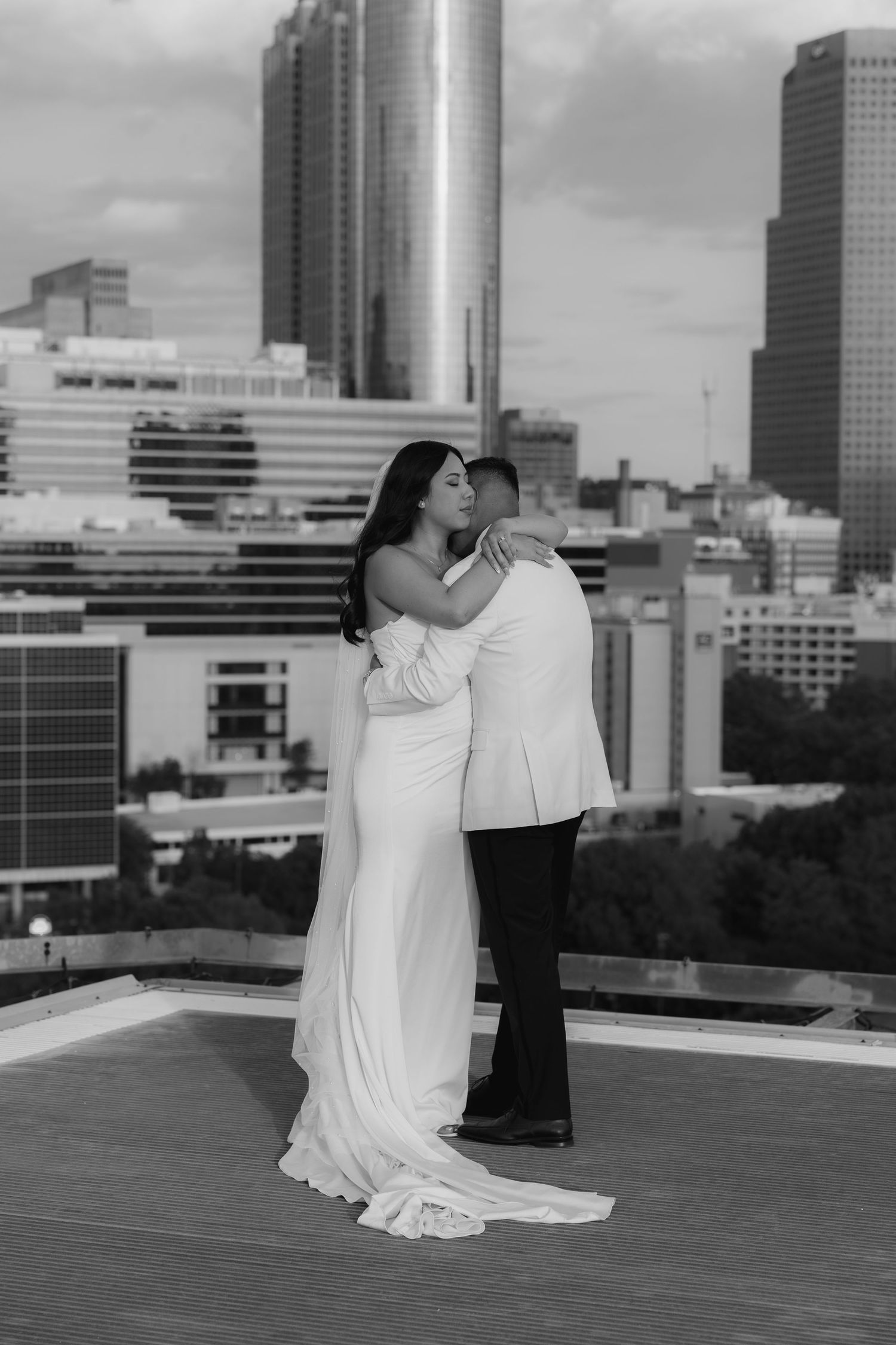 Black and white photo of bride and groom embracing on rooftop with city skyline backdrop.