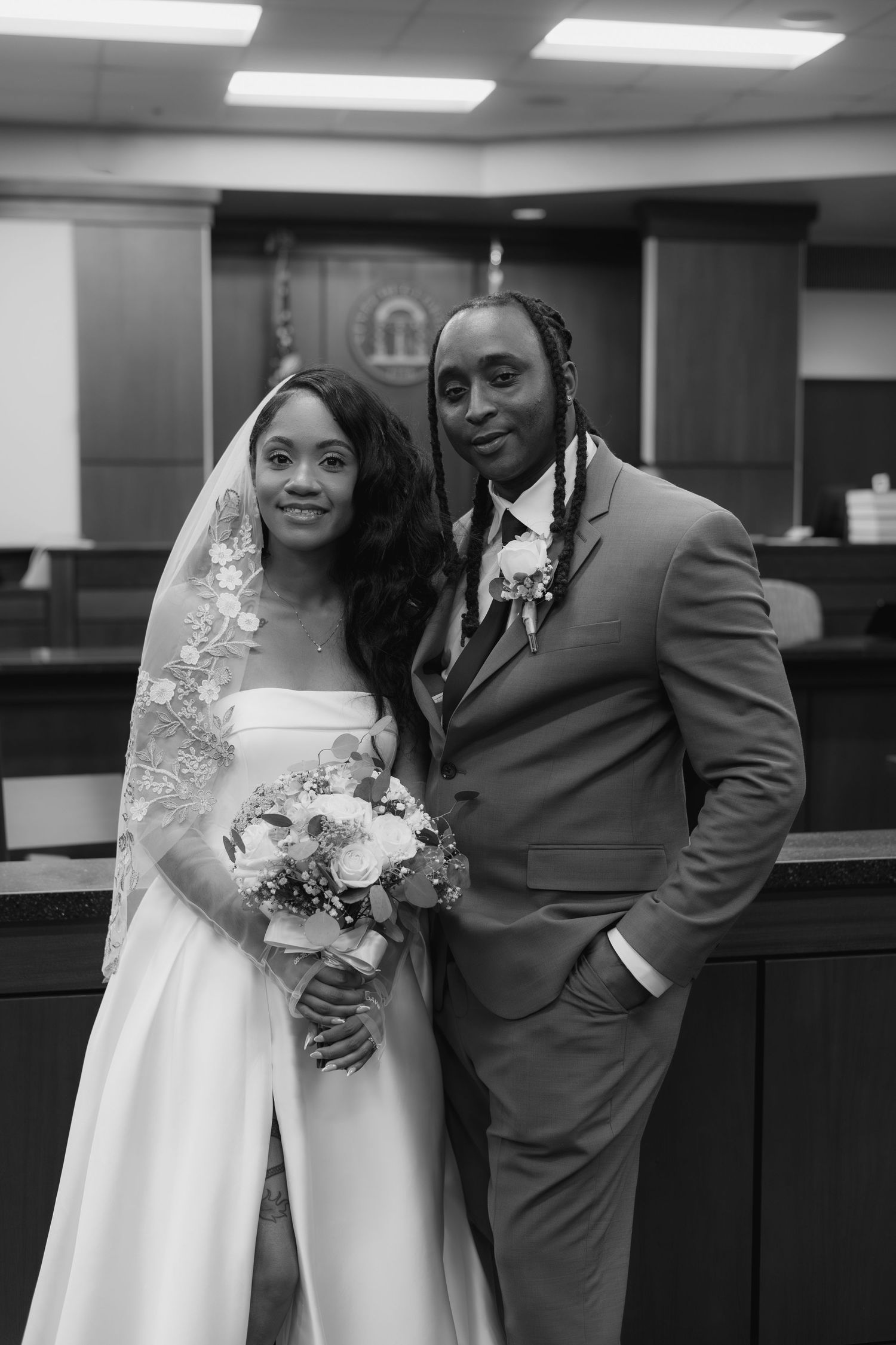 Black and white wedding photo of a smiling couple in a courthouse, bride in white gown with veil, groom in suit with boutonniere.