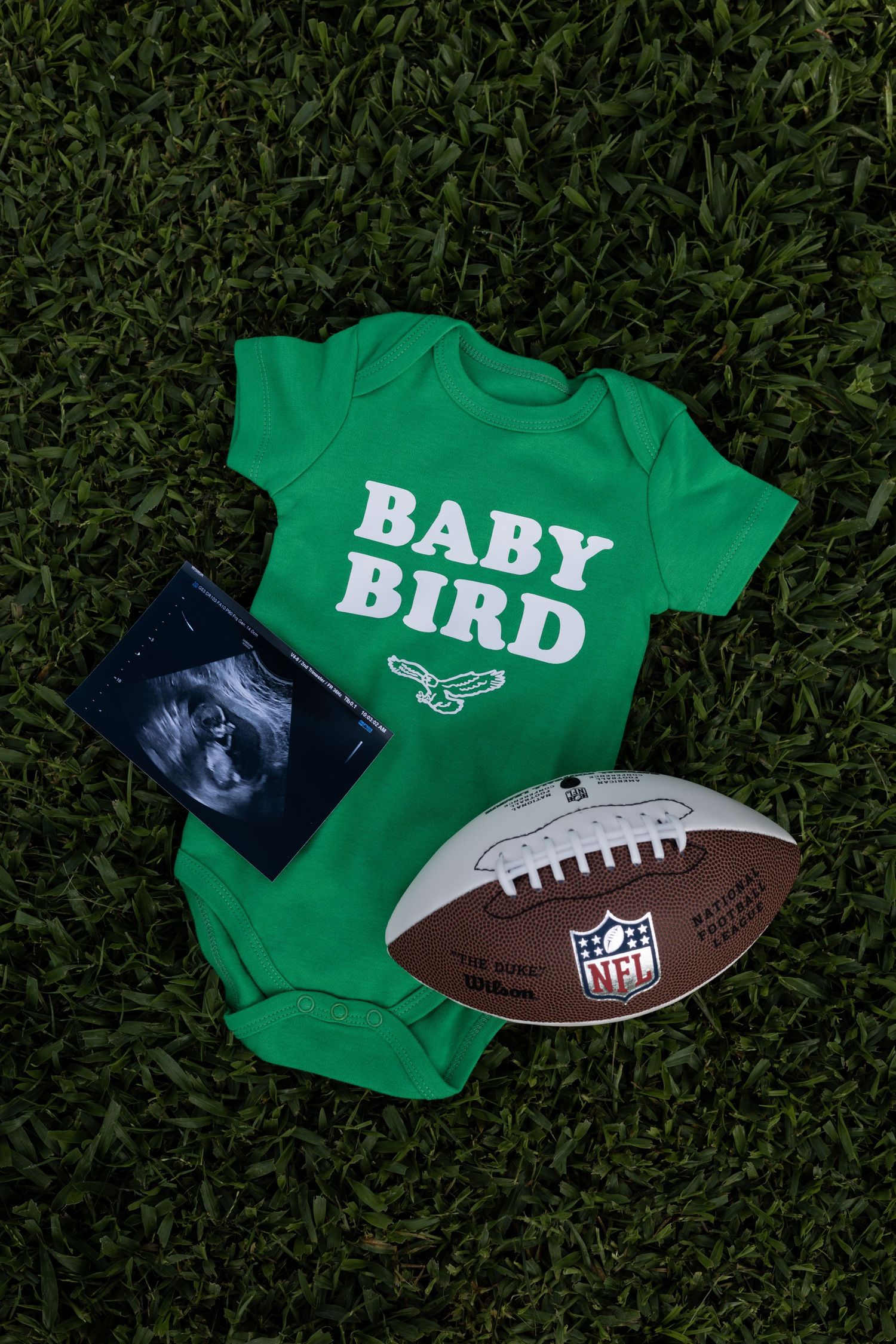 Green baby onesie with 'BABY BIRD' text displayed with NFL football on grass background.