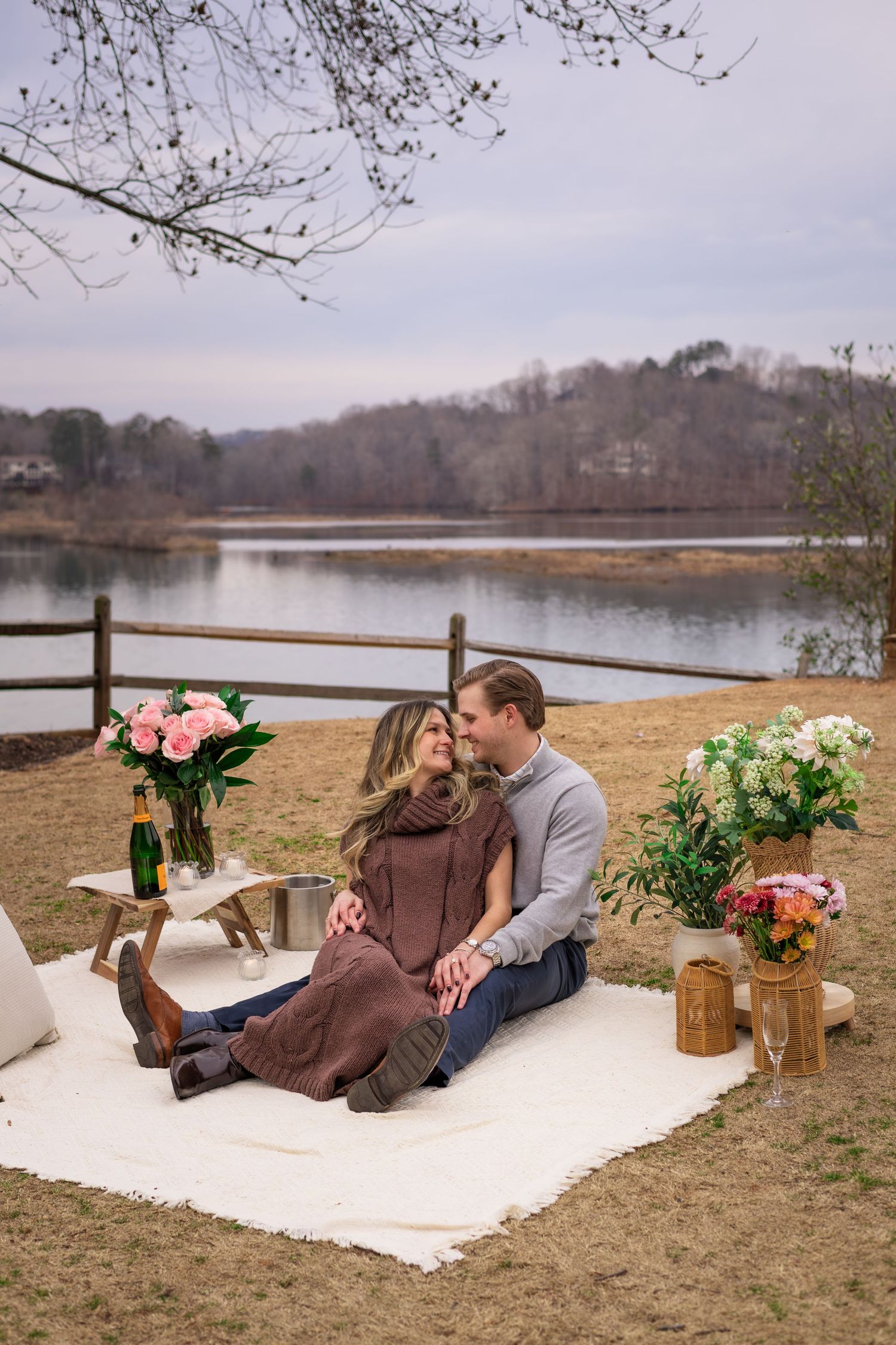 A couple sitting on a blanket in a grassy area by a lake, surrounded by flowers and trees.