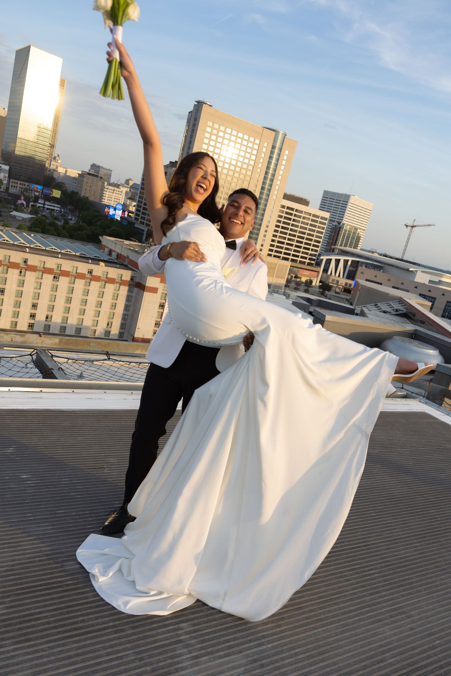 Joyful groom lifting bride in white gown on rooftop with city skyline at sunset.