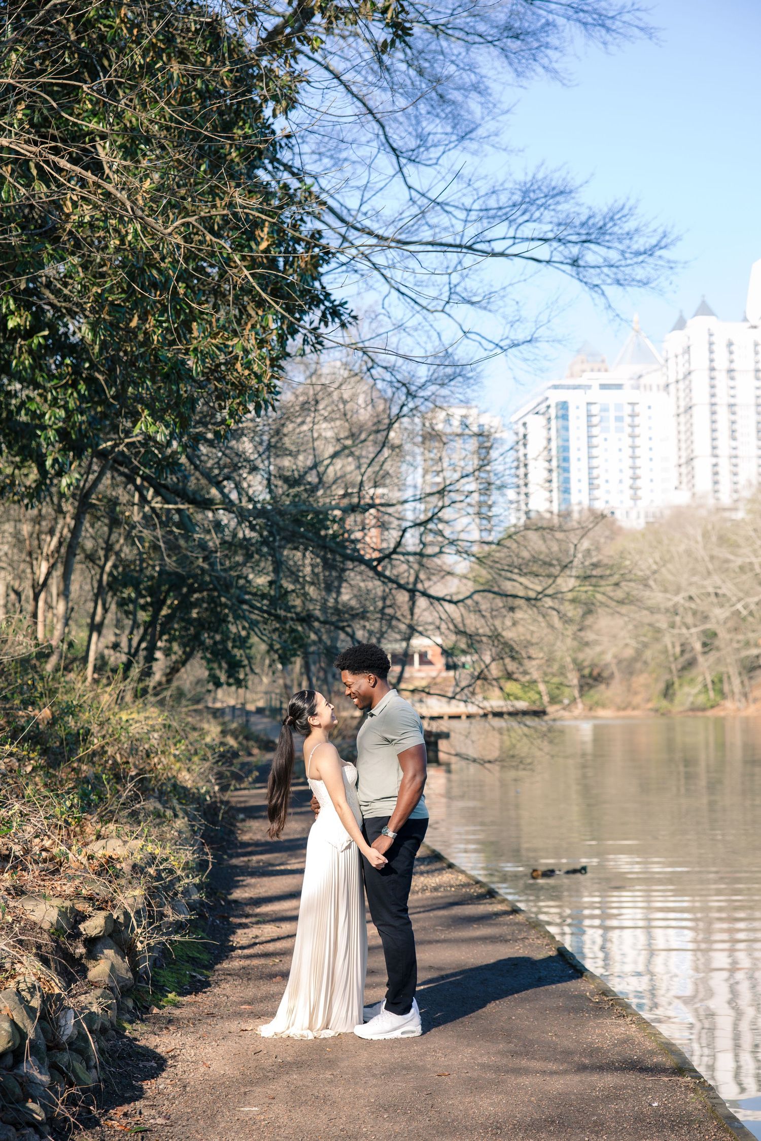 A couple stands together on a lakeside path with tall buildings visible in the distance across the water.