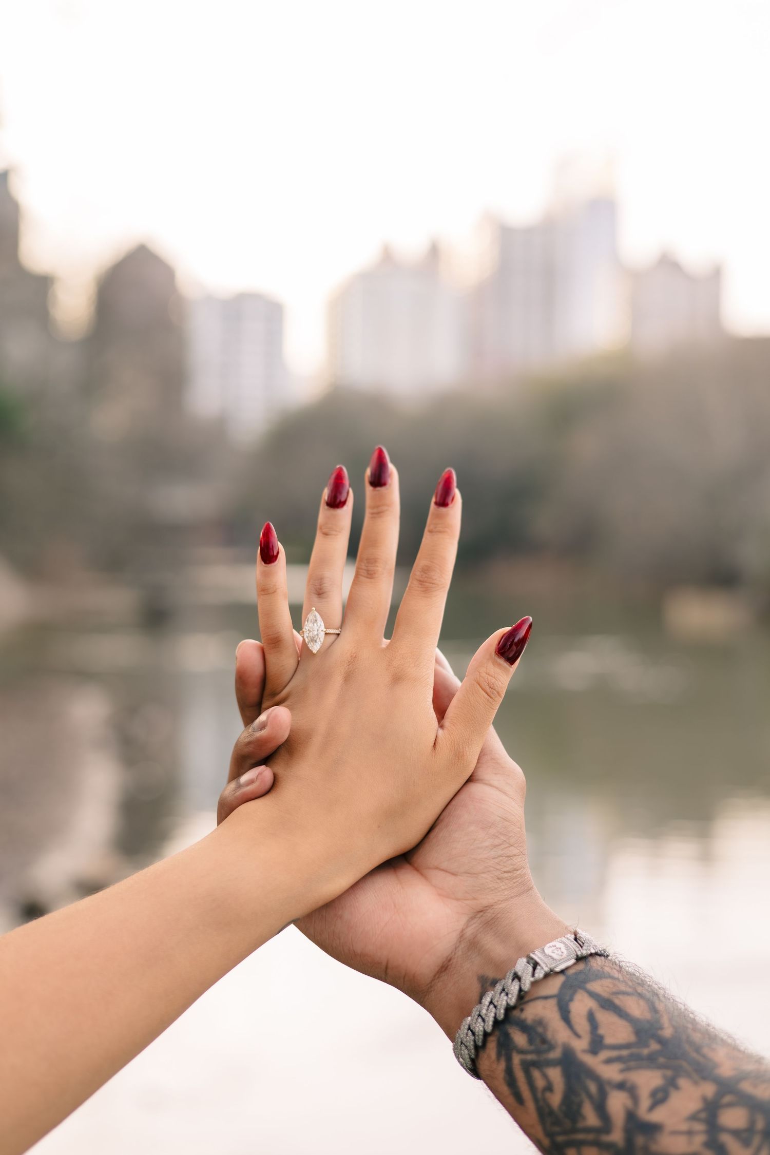 Two hands touch with one showing red manicured nails and diamond ring against a blurred cityscape and lake background.