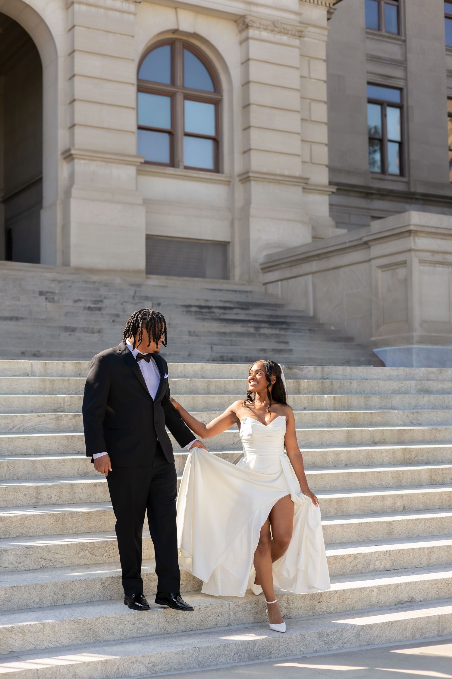 A bride and groom walk down grand marble steps outside a classical building, she lifts her white gown while smiling at him.