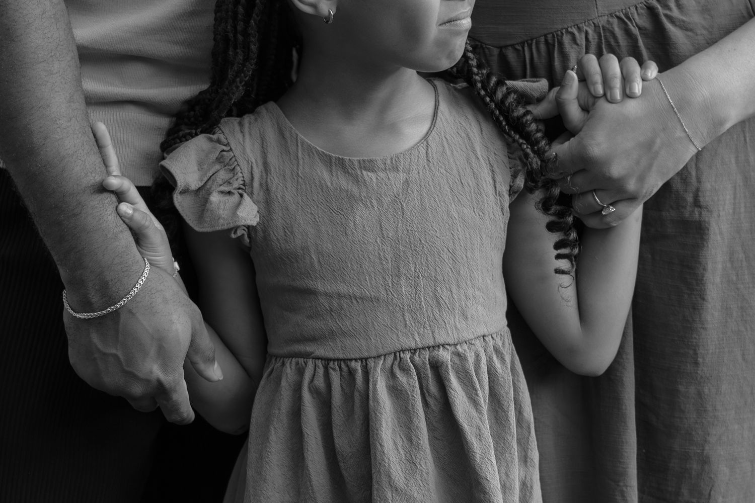 Black and white photo of a young girl in a dress holding hands with two adults on either side of her.